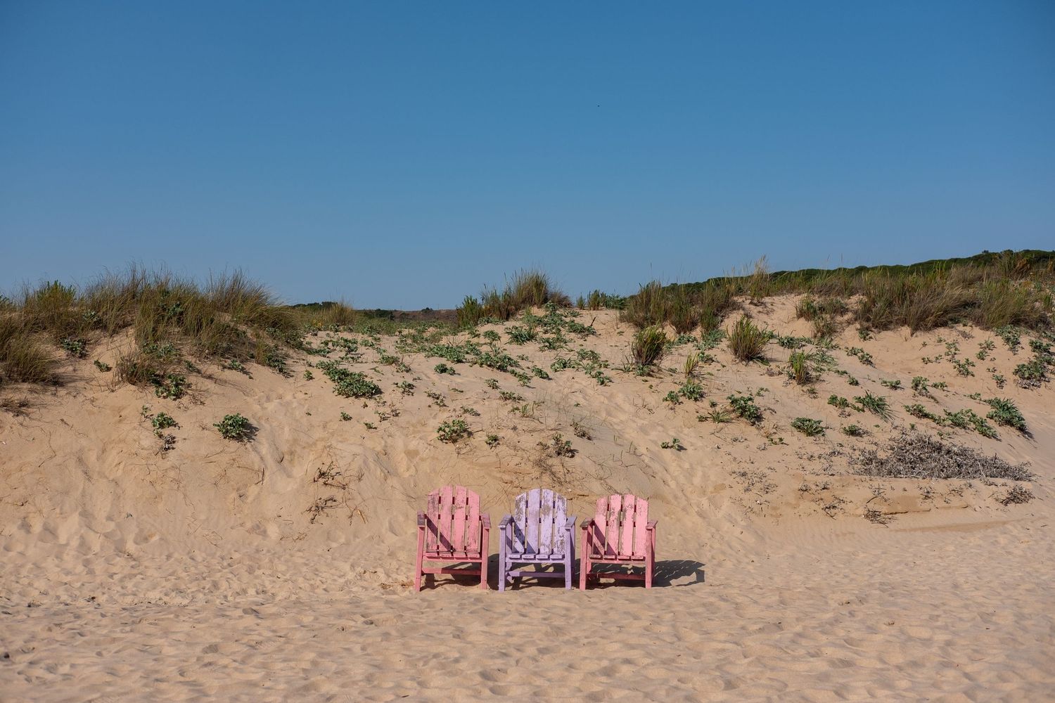 Two pink beach chairs sitting empty on sandy dunes under clear blue sky.