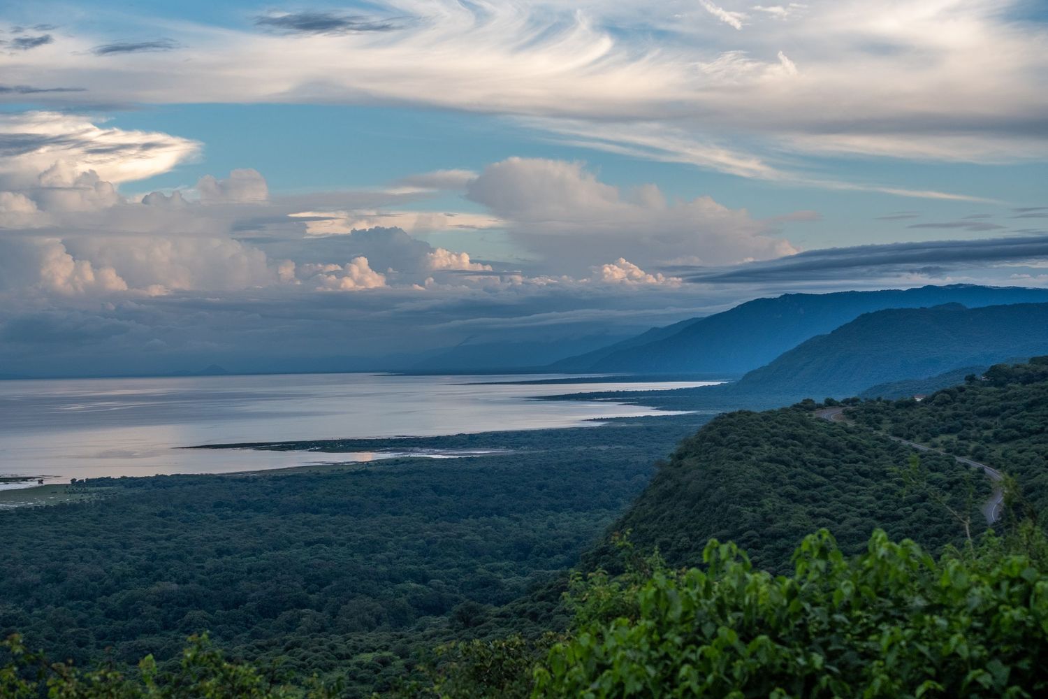 Sweeping coastal vista with mountains and clouds reflecting on calm waters at sunset.
