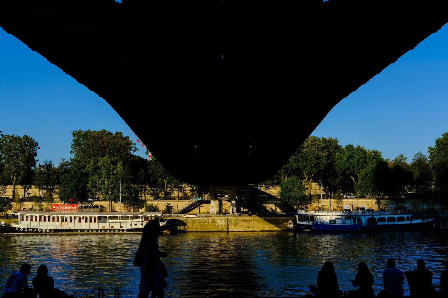 Silhouetted bridge view overlooking boats docked along a sunny riverbank.