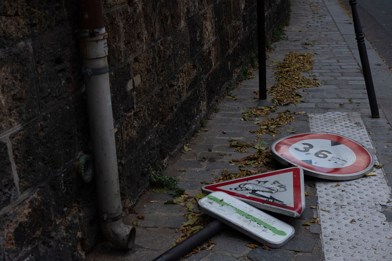 Fallen traffic signs lean against weathered stone wall on cobblestone sidewalk.