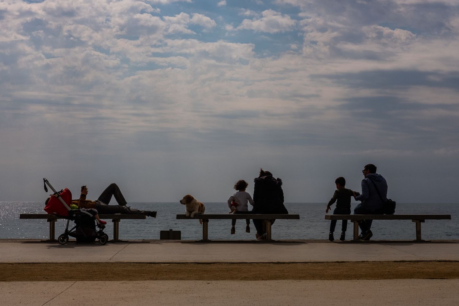 Silhouettes of people sitting on benches against a cloudy sky at sunset.
