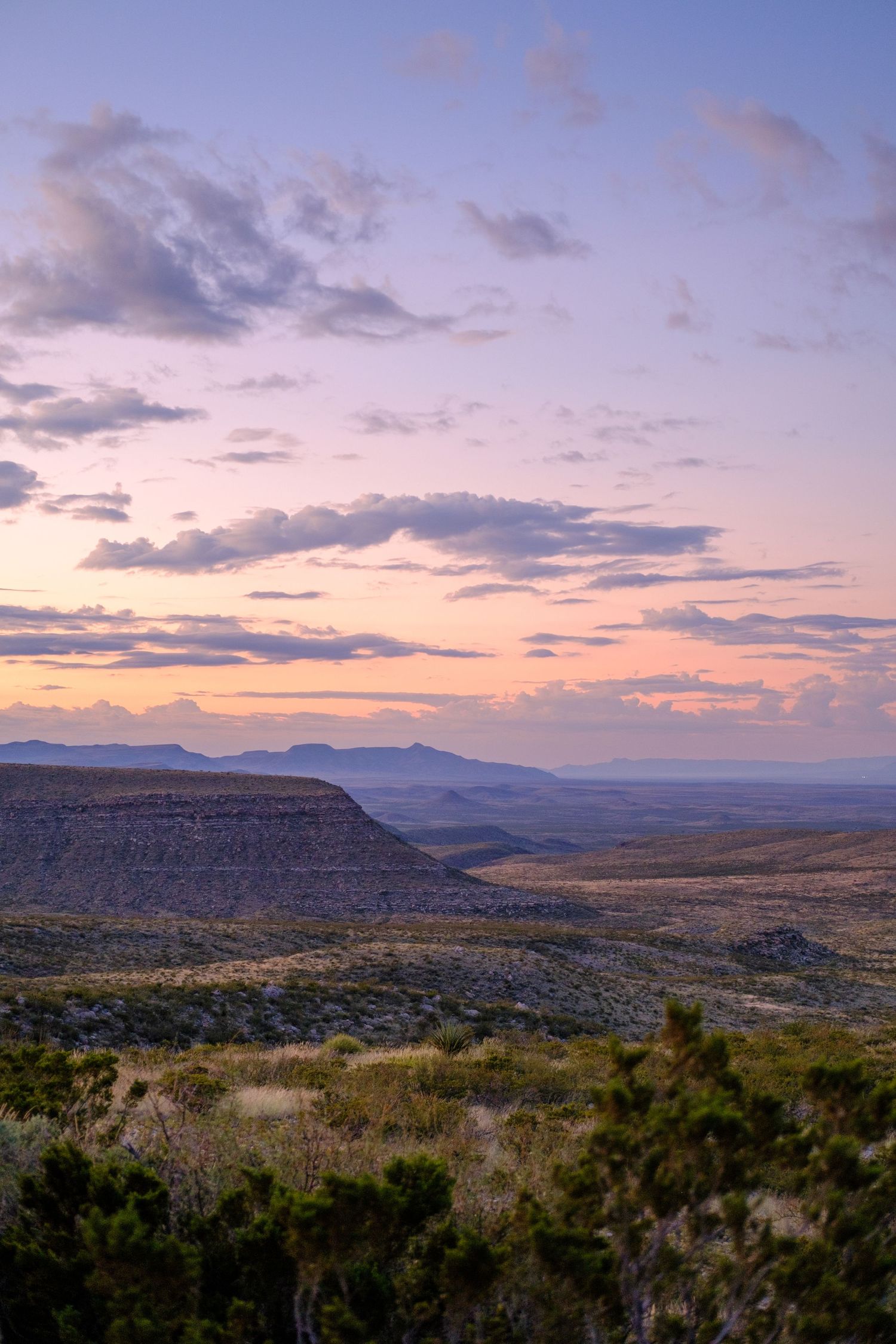 Panoramic sunset view over rugged mountains and valleys with scattered vegetation in warm evening light.