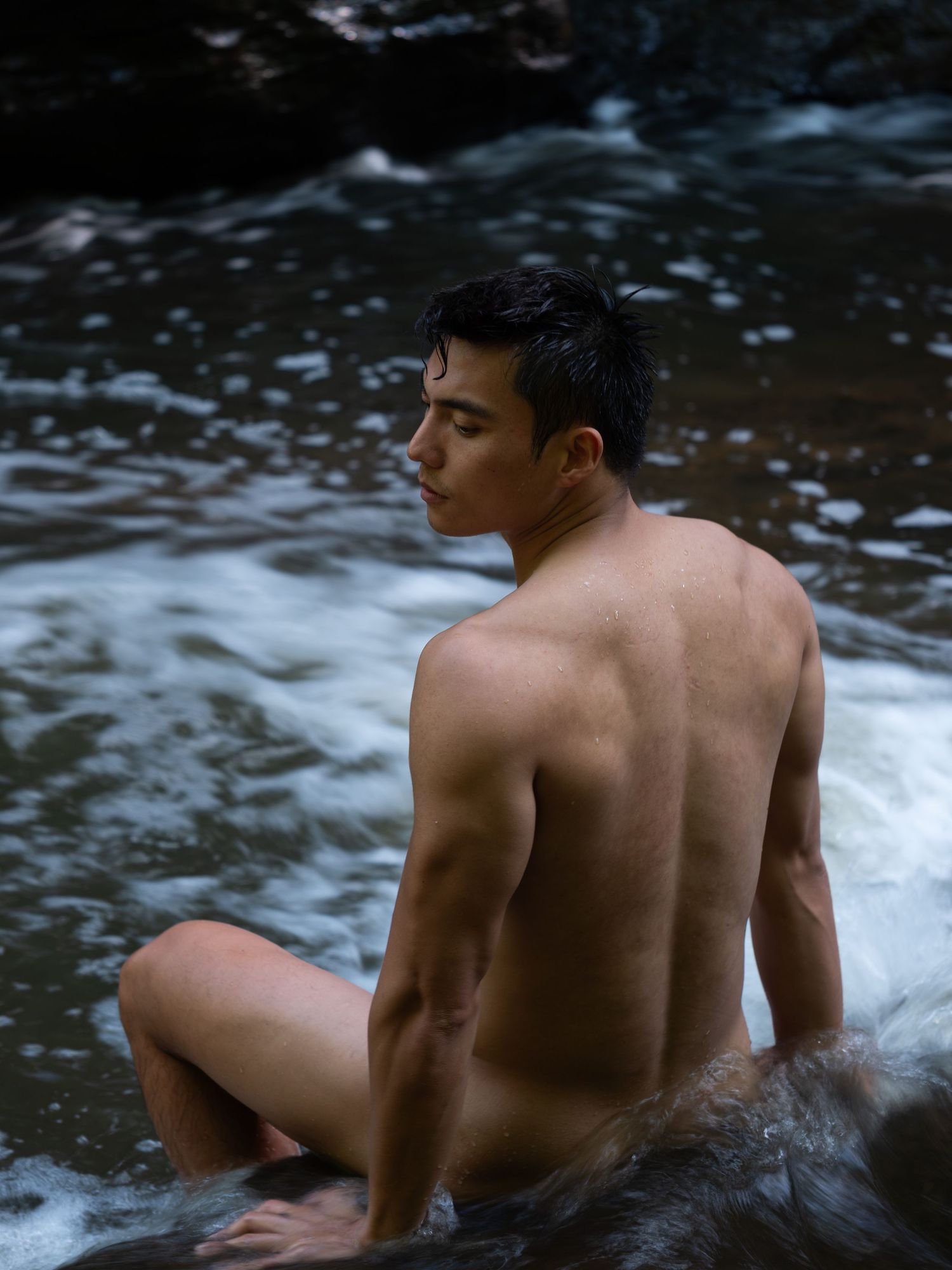 Shirtless figure sitting in flowing water against dark rocky background at dusk.