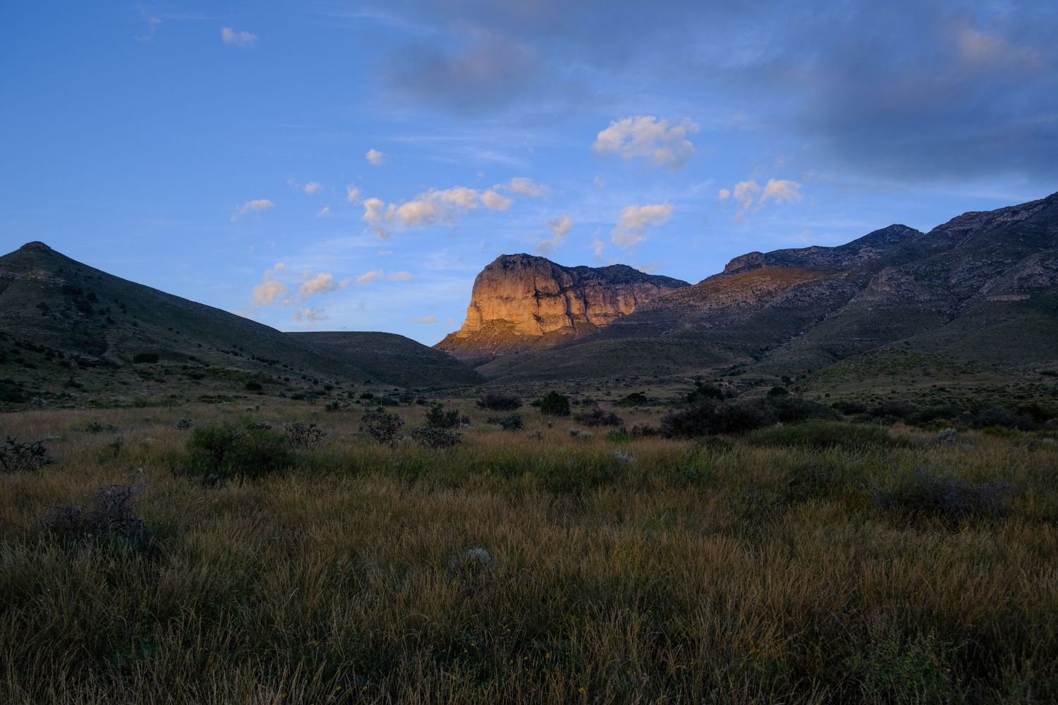 Dramatic sunset light illuminates a towering cliff face in a rugged mountain landscape with desert scrubland in the foreground.