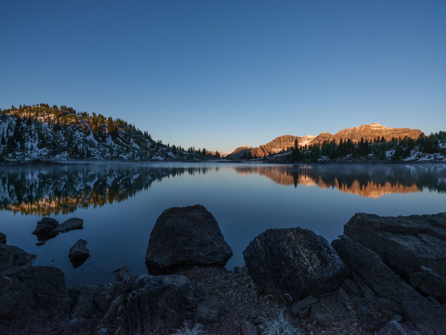 Tranquil mountain lake at sunset reflects snow-capped peaks and rocky shoreline in panoramic view.