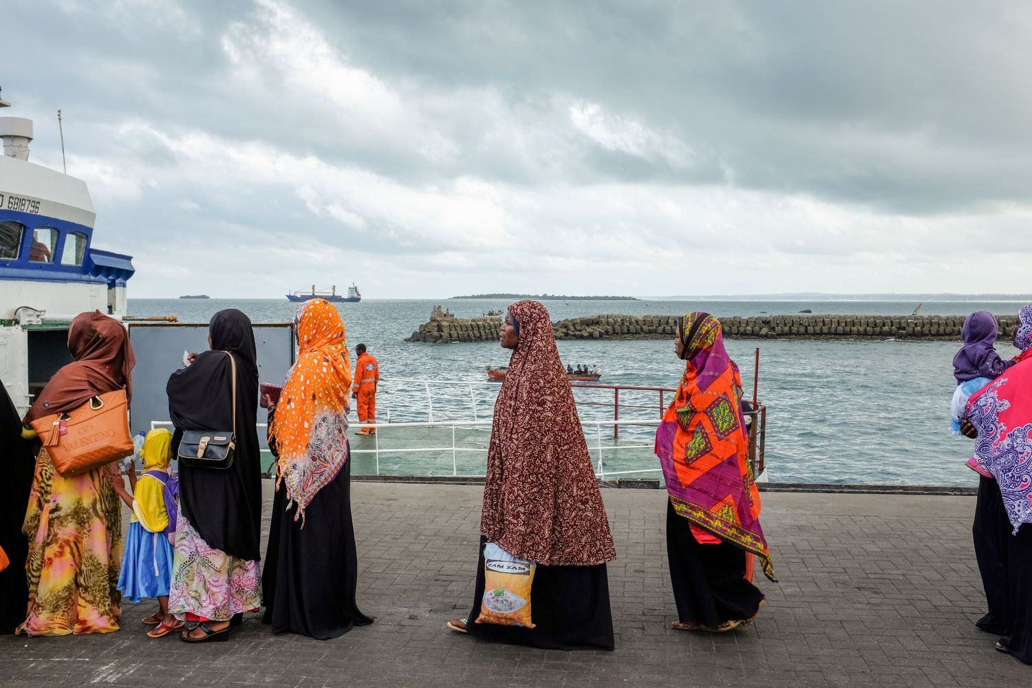 Colorfully dressed people walk along a beach with waves and cloudy skies in the background.