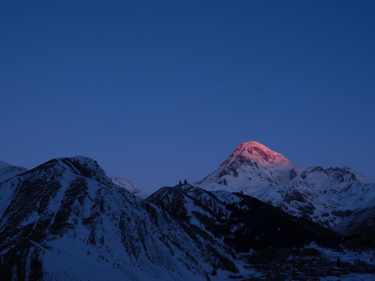 Snow-capped mountain peak glowing pink at sunset against a deep blue sky.