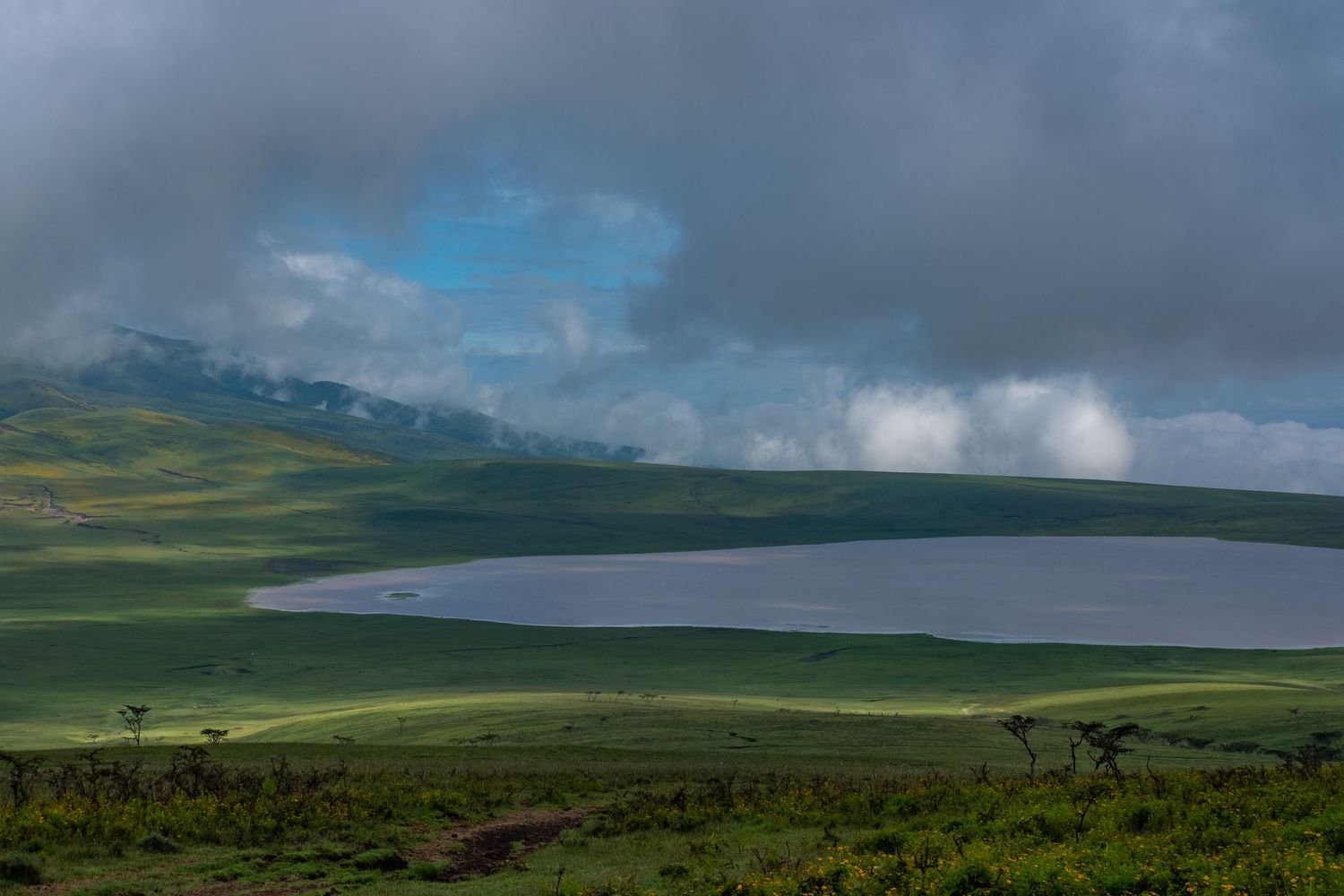 Panoramic view of a lake surrounded by lush green hills and cloudy mountain peaks.