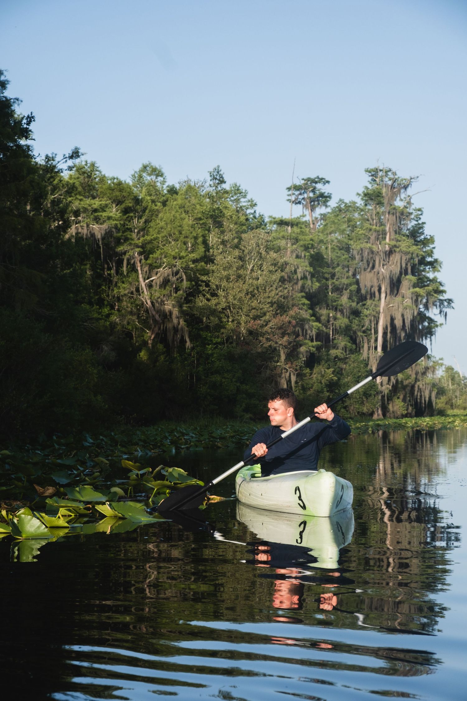 Kayaker paddles through cypress swamp surrounded by lily pads and Spanish moss.