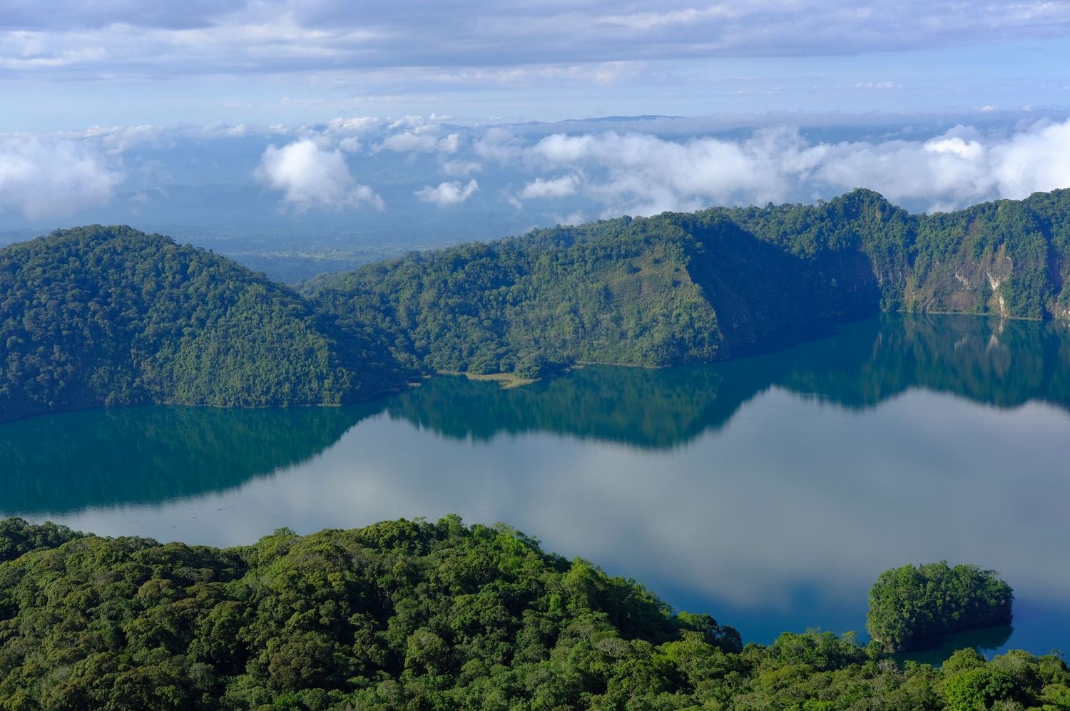 Panoramic view of a pristine lake surrounded by lush green mountains under blue skies.