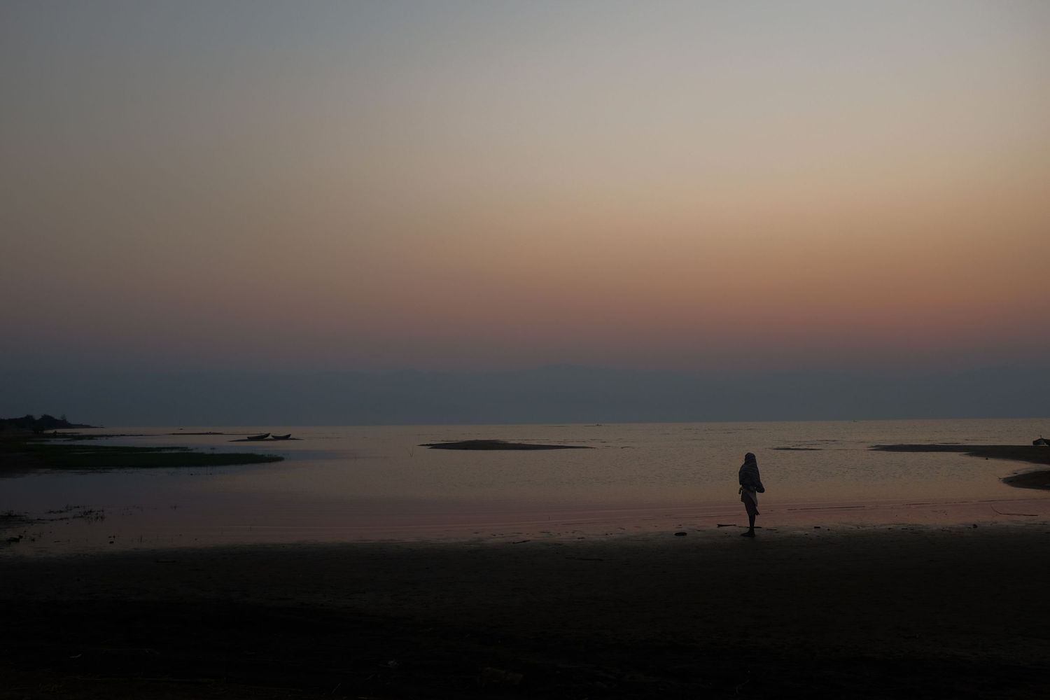 Silhouetted fishing boats line the shoreline at sunset creating a peaceful coastal scene.