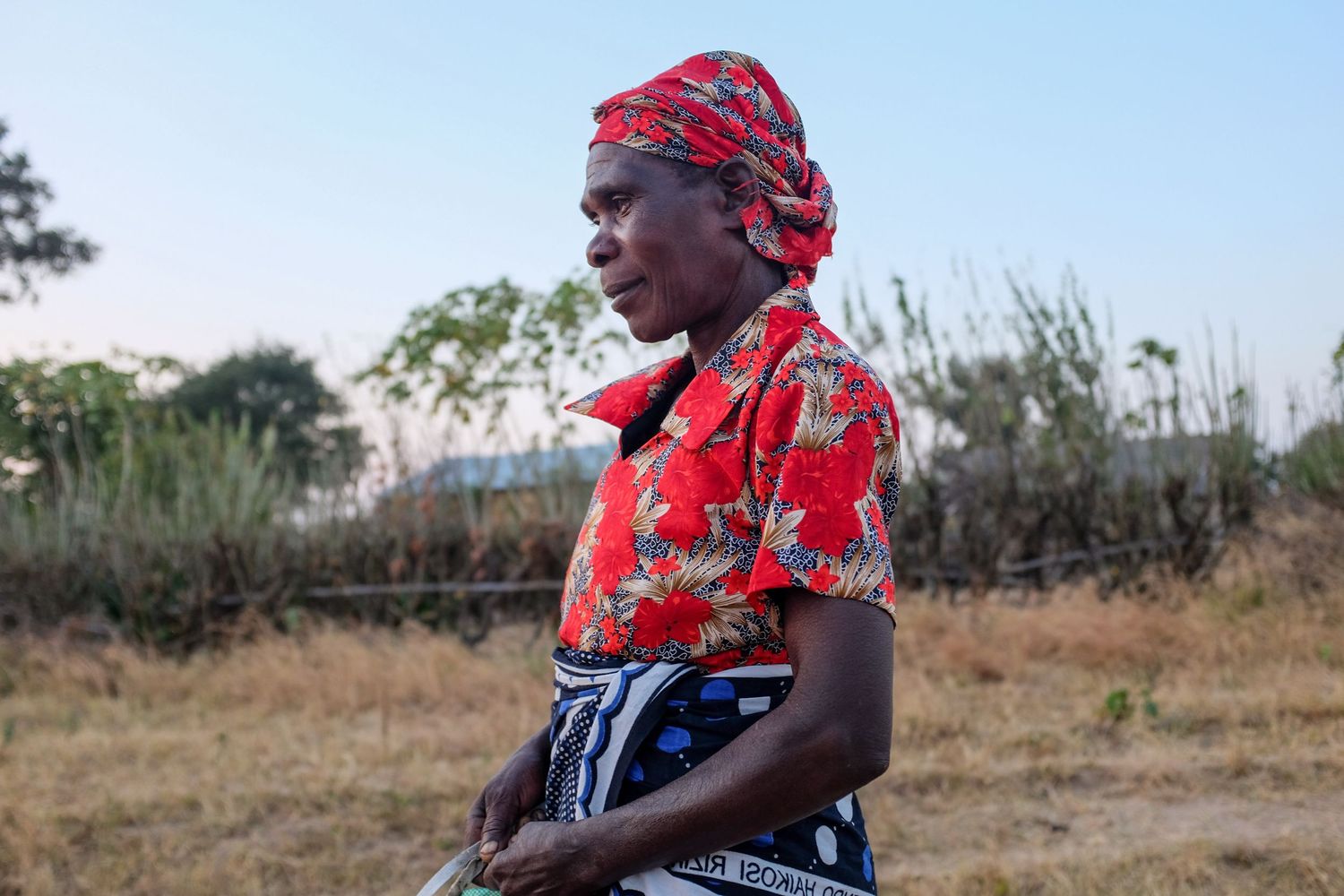 A person wearing a red headscarf and traditional dress carries a basket while walking through a rural field.