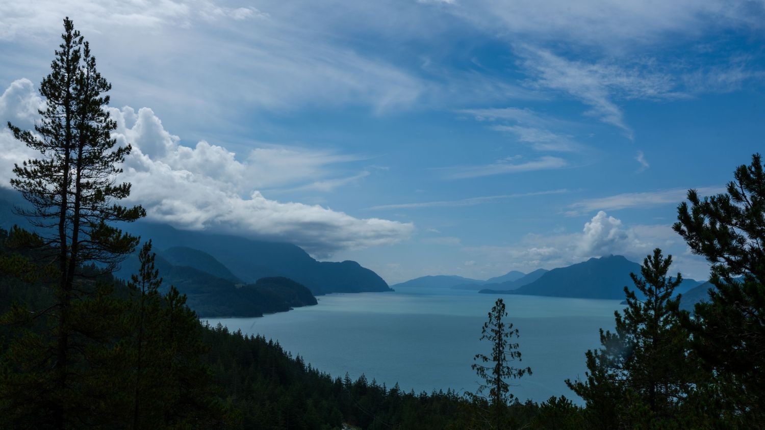 Panoramic mountain lake view with evergreen trees and dramatic clouds against blue sky.