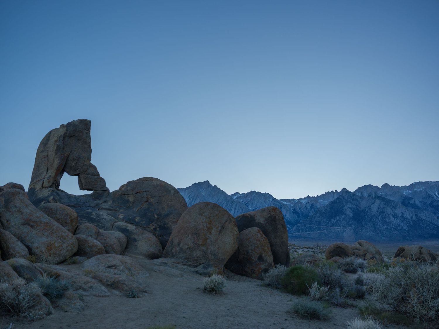 Rocky desert formations and mountains under a deep blue sky at dusk.