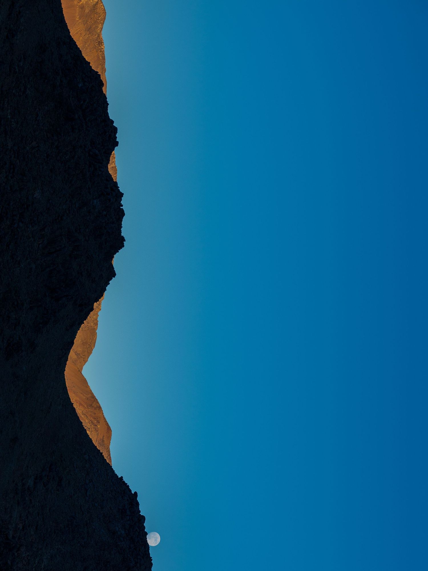 Rocky mountain silhouette against a bright blue sky with a small white moon visible.