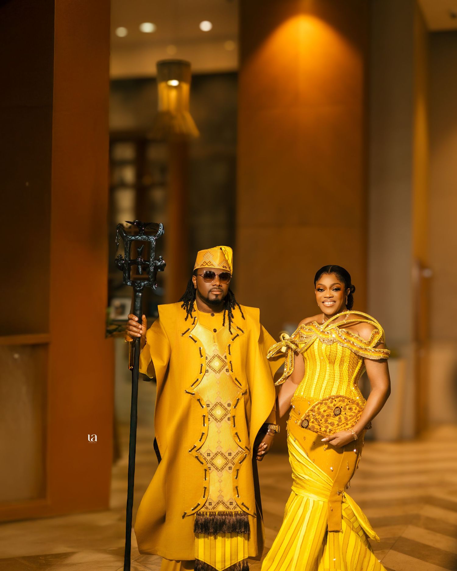 A couple in matching yellow and gold traditional formal wear poses in an elegant hallway.