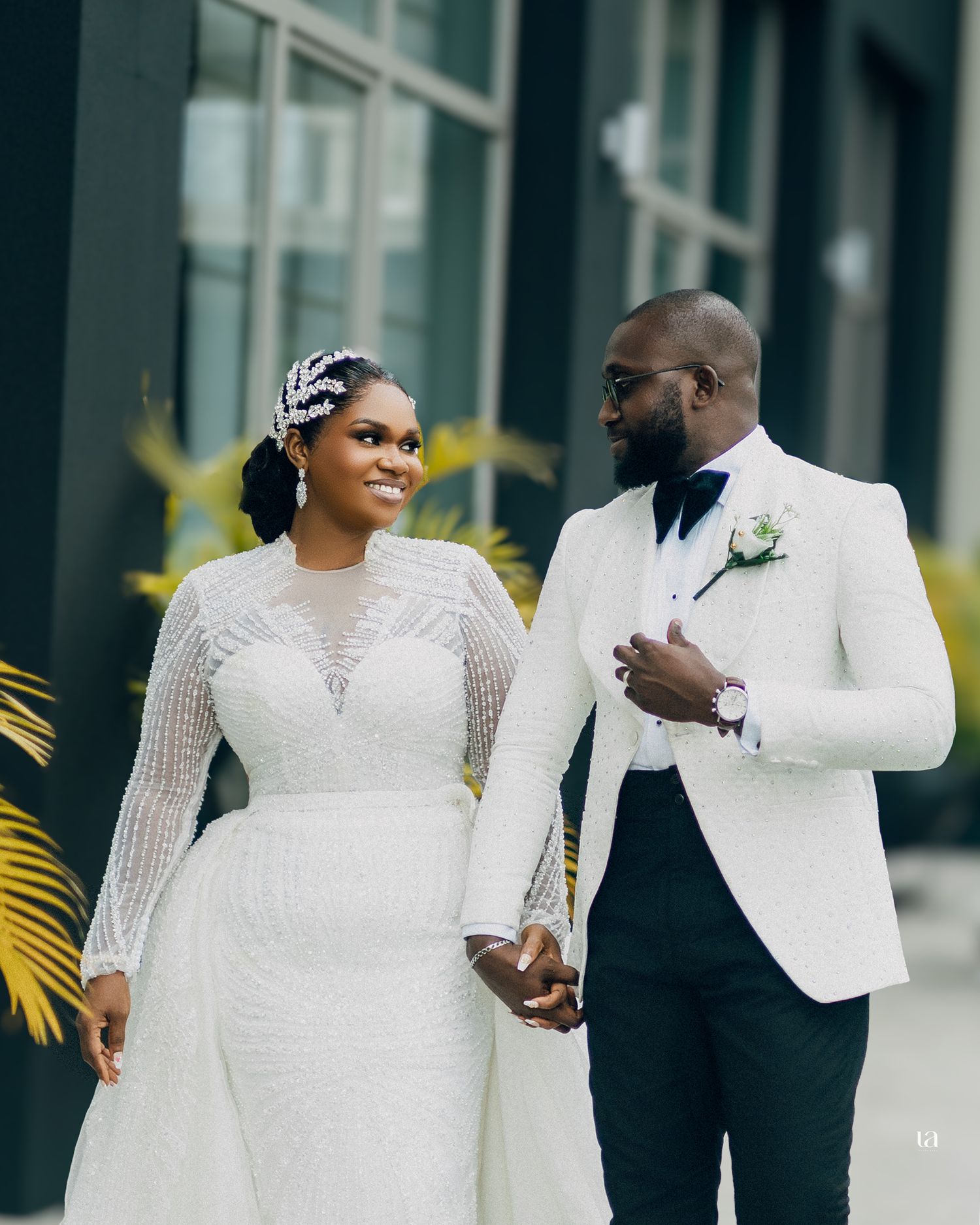 Wedding party in elegant white attire poses together outside modern glass building with palm fronds visible.