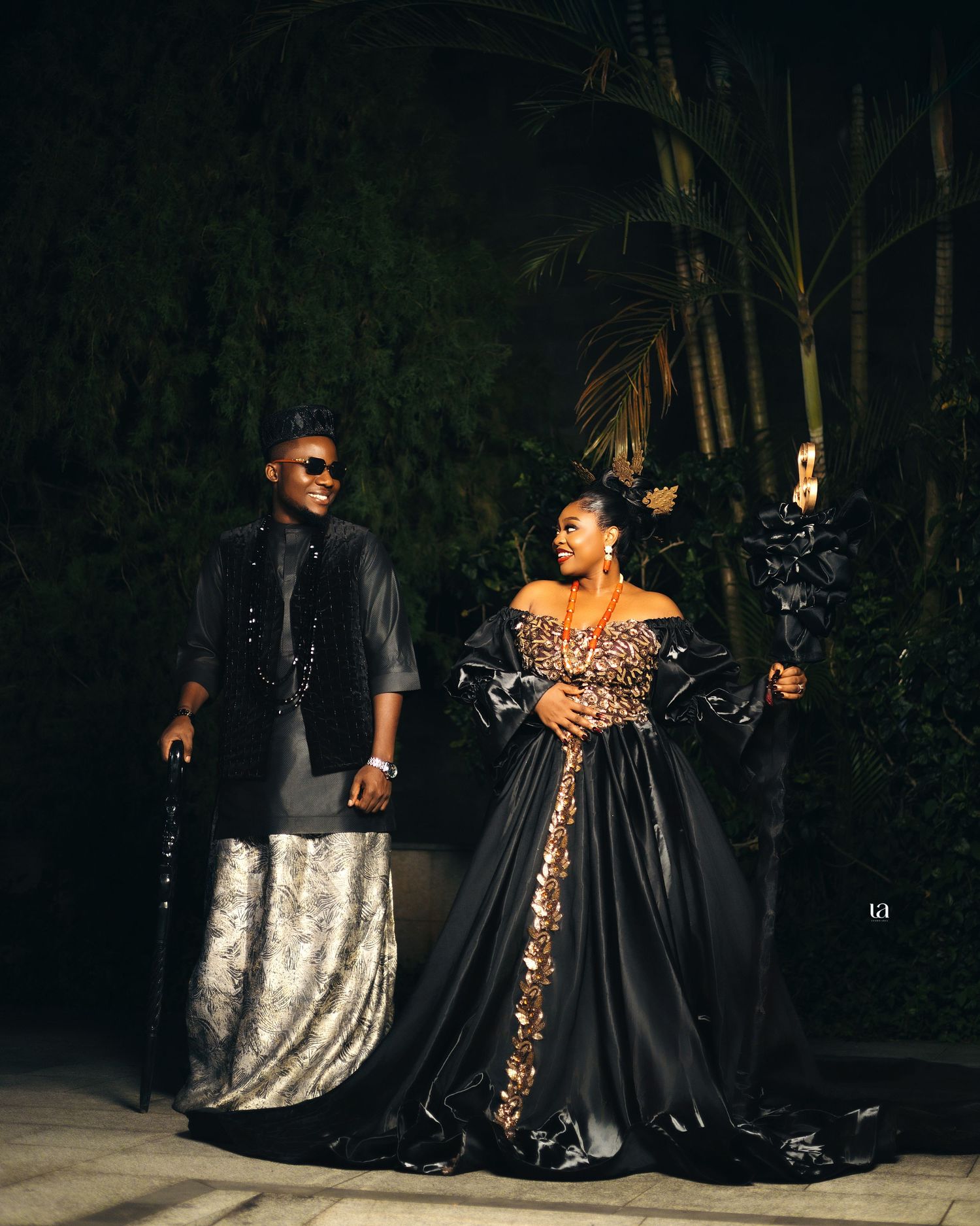 Dramatic outdoor photoshoot featuring elegant couple in flowing black and gold evening attire.