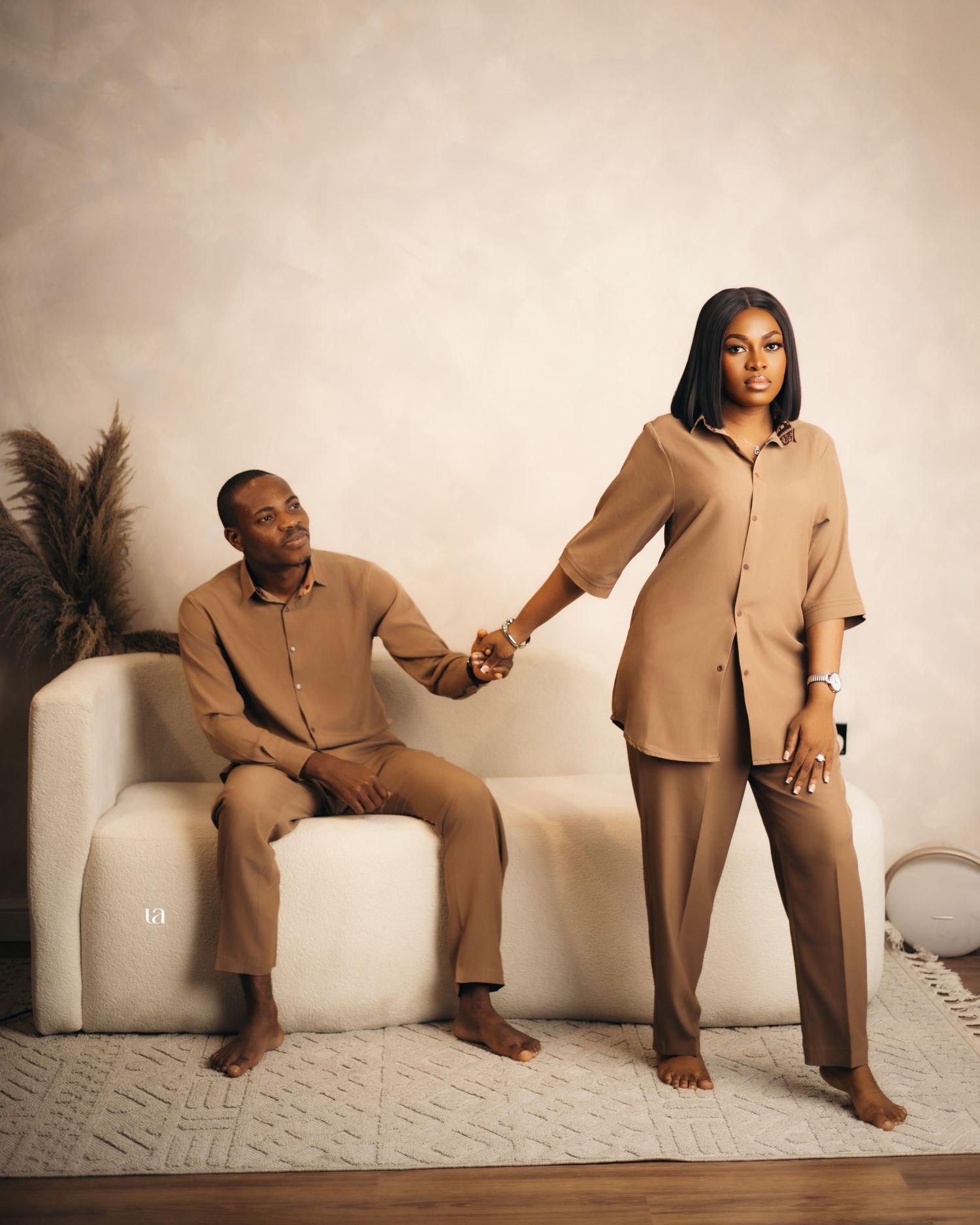 Playful couple's photoshoot featuring relaxed poses on white couch in matching beige outfits.