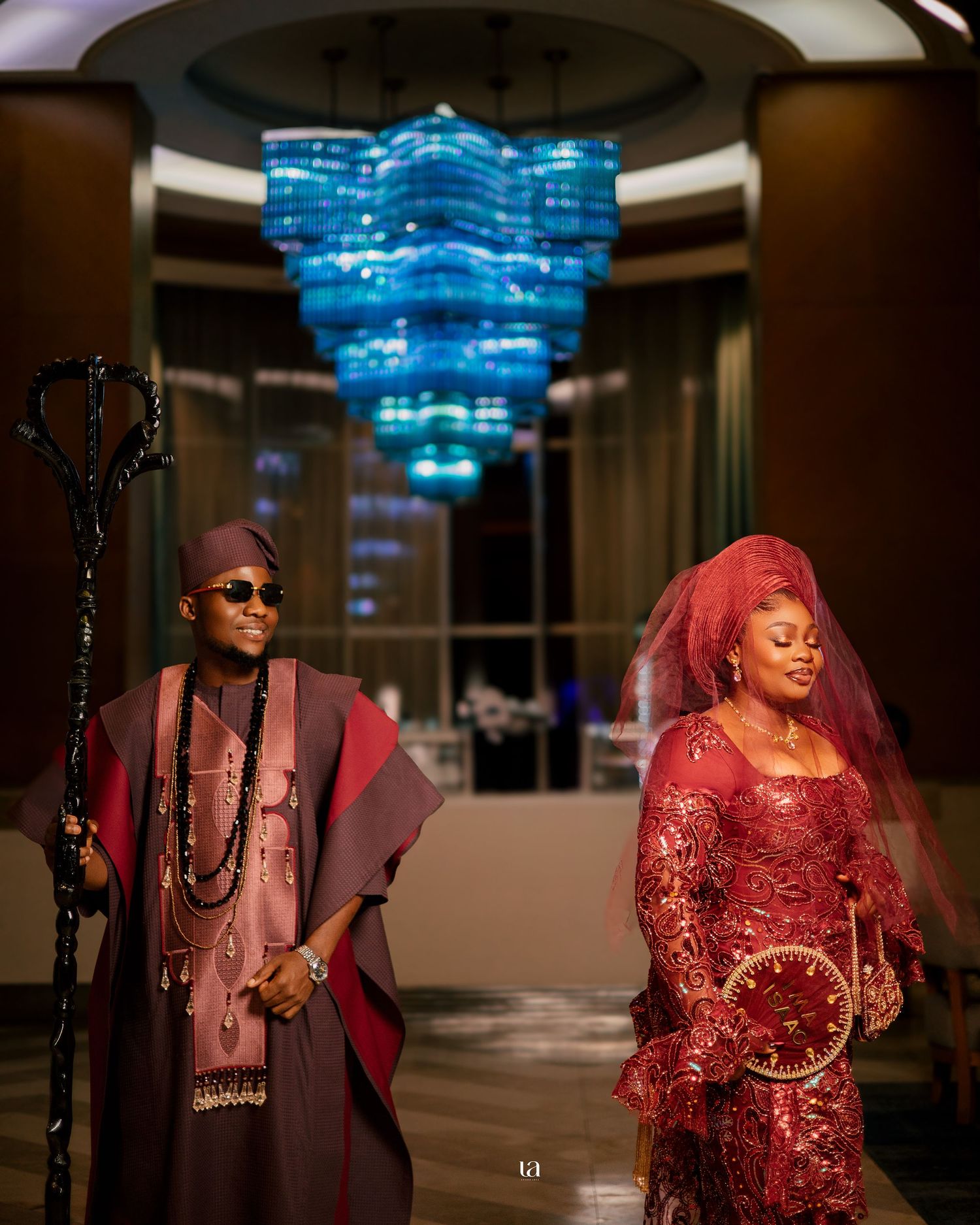 A couple in traditional attire poses under a blue chandelier in formal wear.