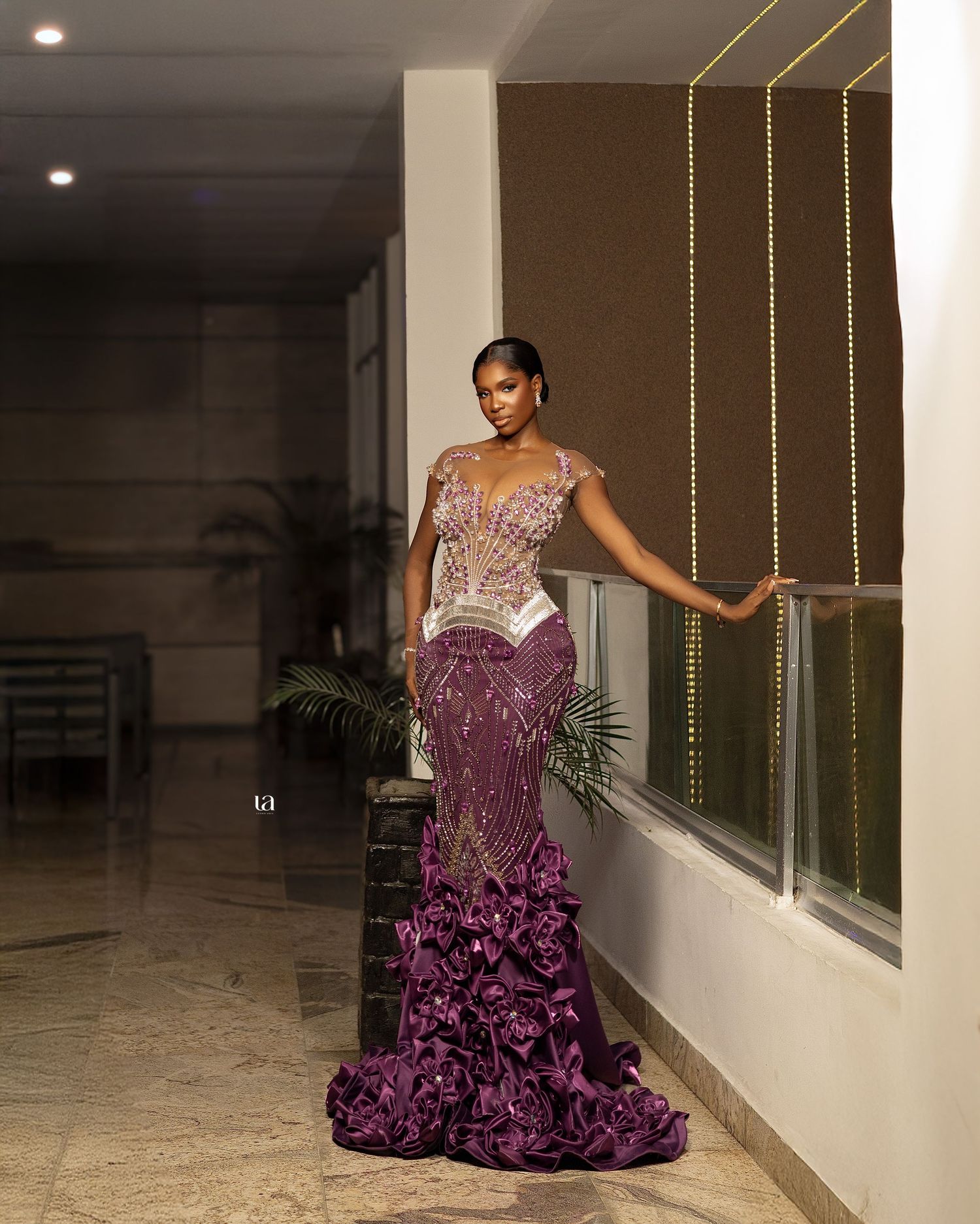 Model in purple embellished evening gown poses in elegant hallway with string lights.