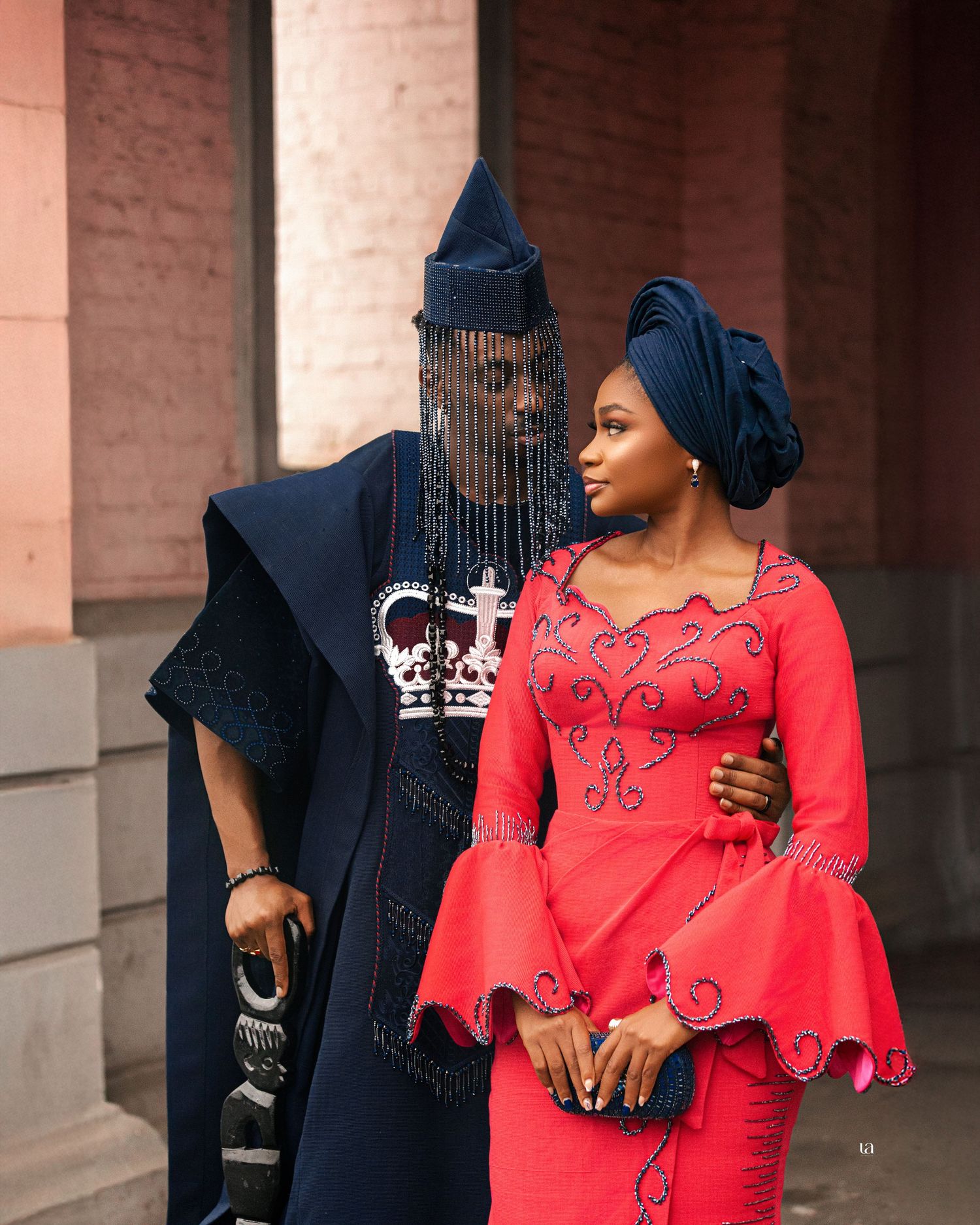 Couple in contrasting red and black formal wear posing under pink brick colonnade.