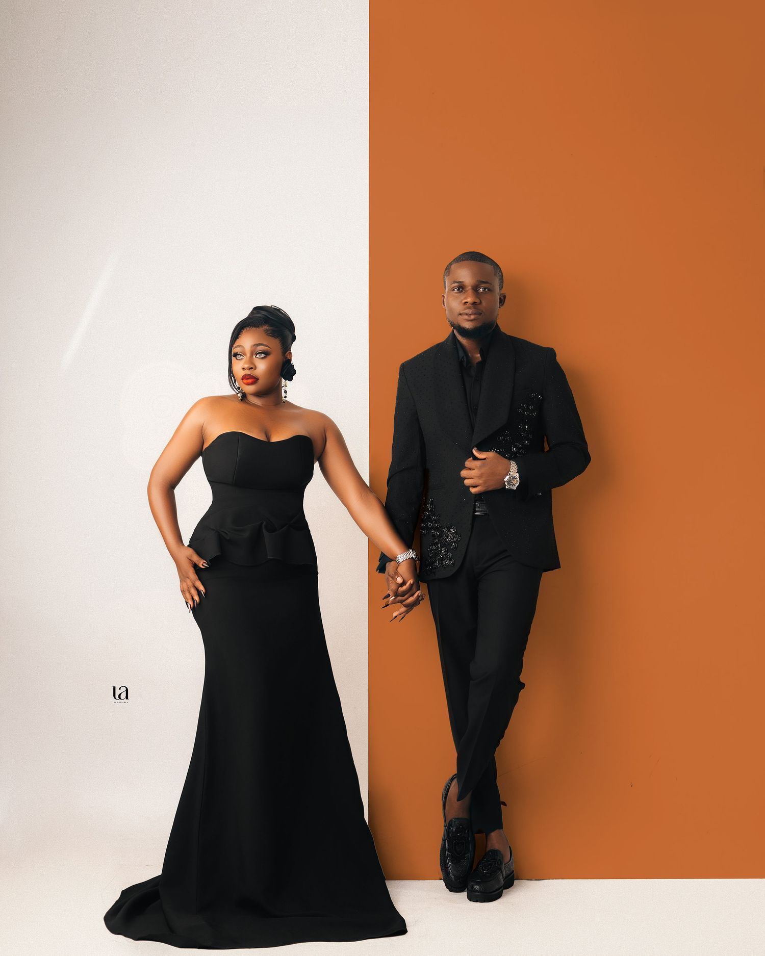 Stylish couple poses against orange and white backdrop wearing coordinated black formal wear.
