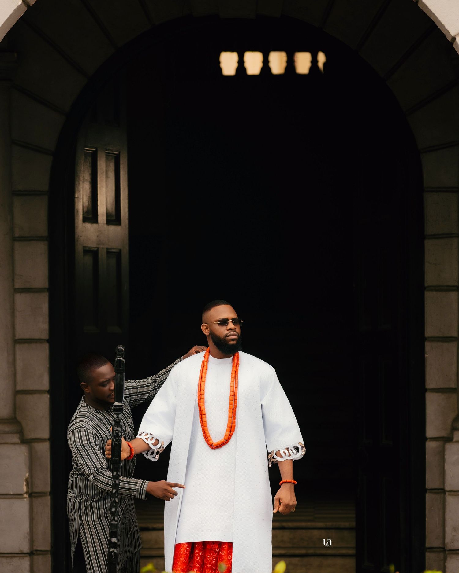 People in traditional white and orange Nigerian attire stand beneath stone archways.