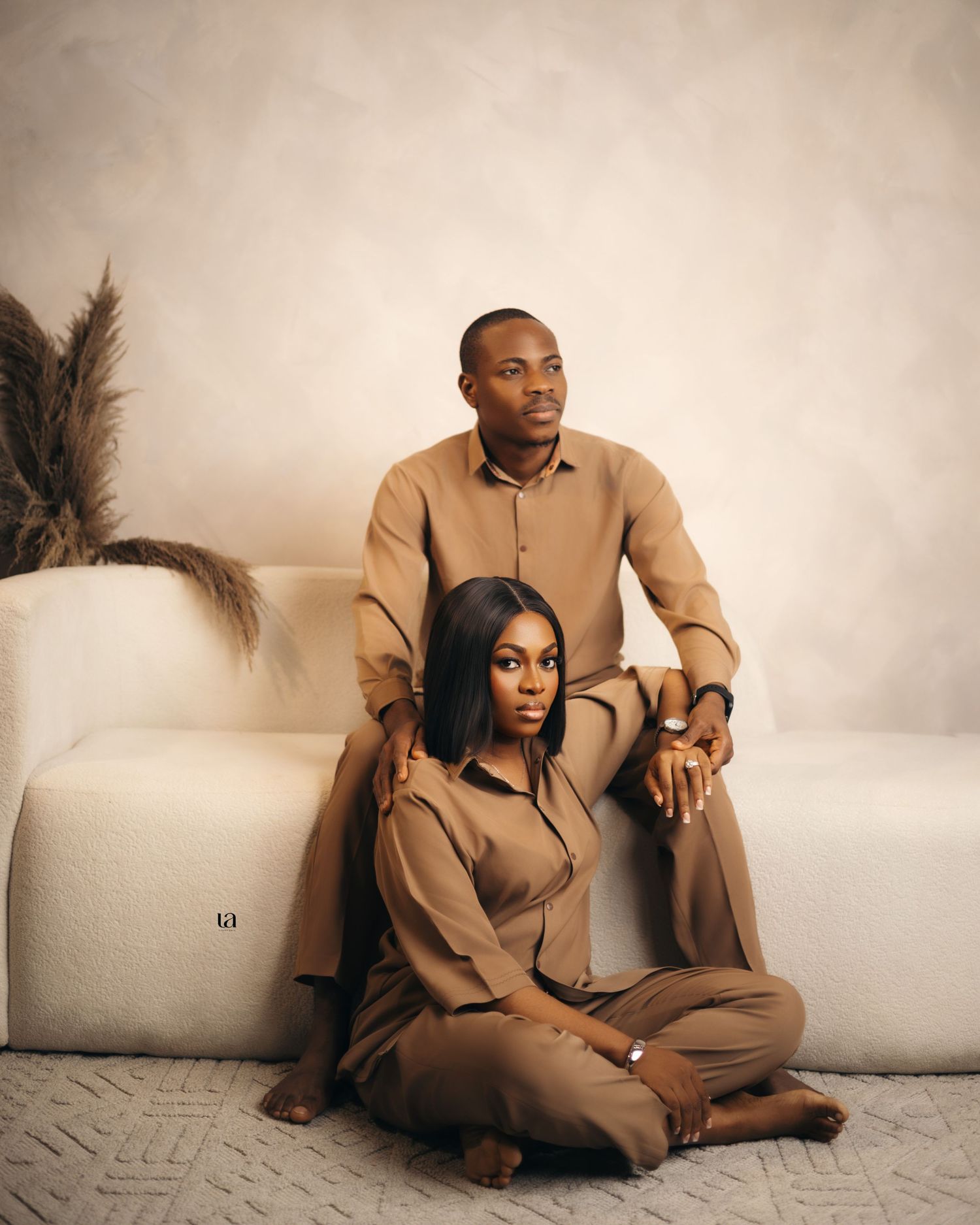 Couple lounges on white sofa in matching tan suits during contemporary photo session.