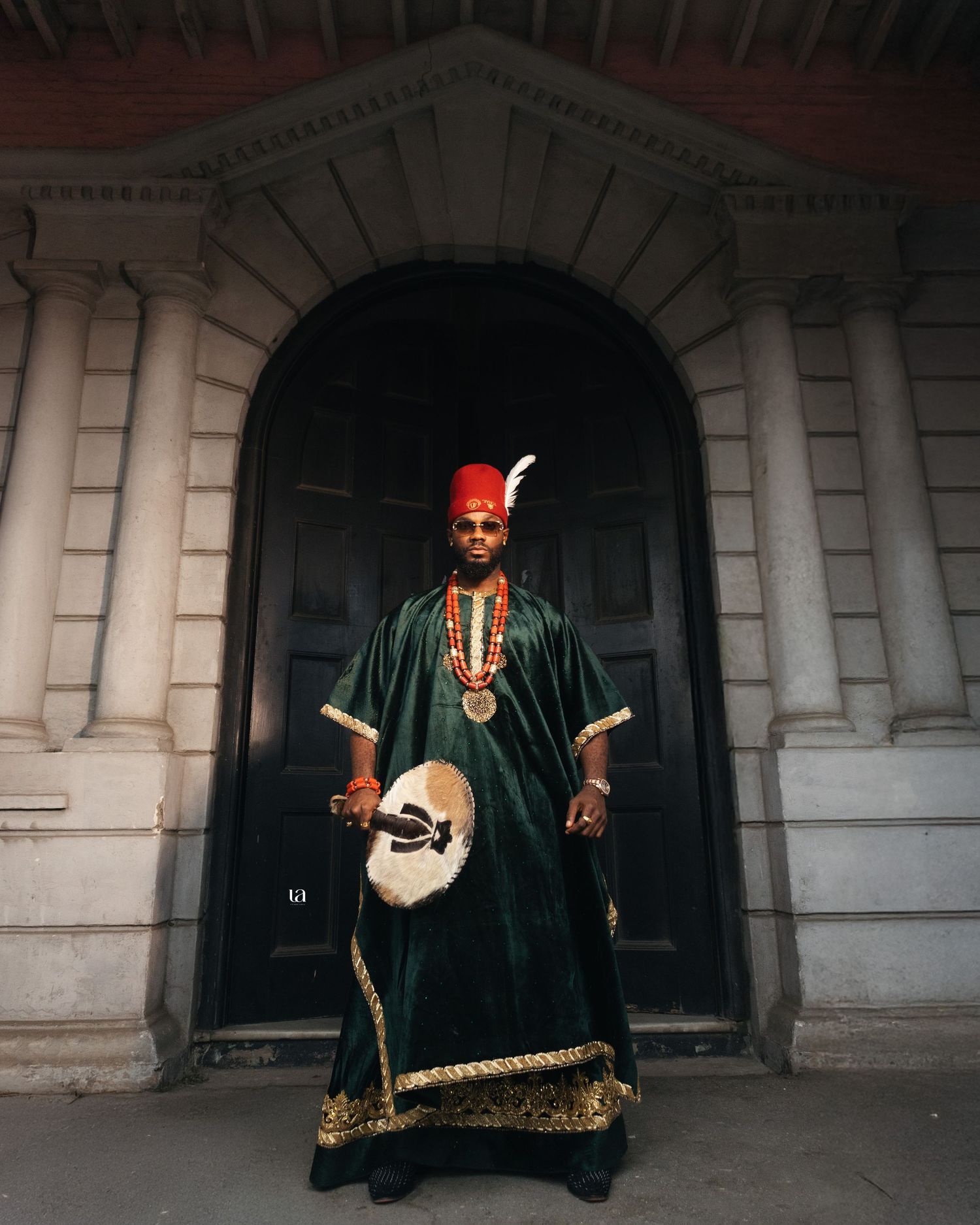 Traditional Nigerian man in dark robe and red cap poses in arched doorway of historic building.