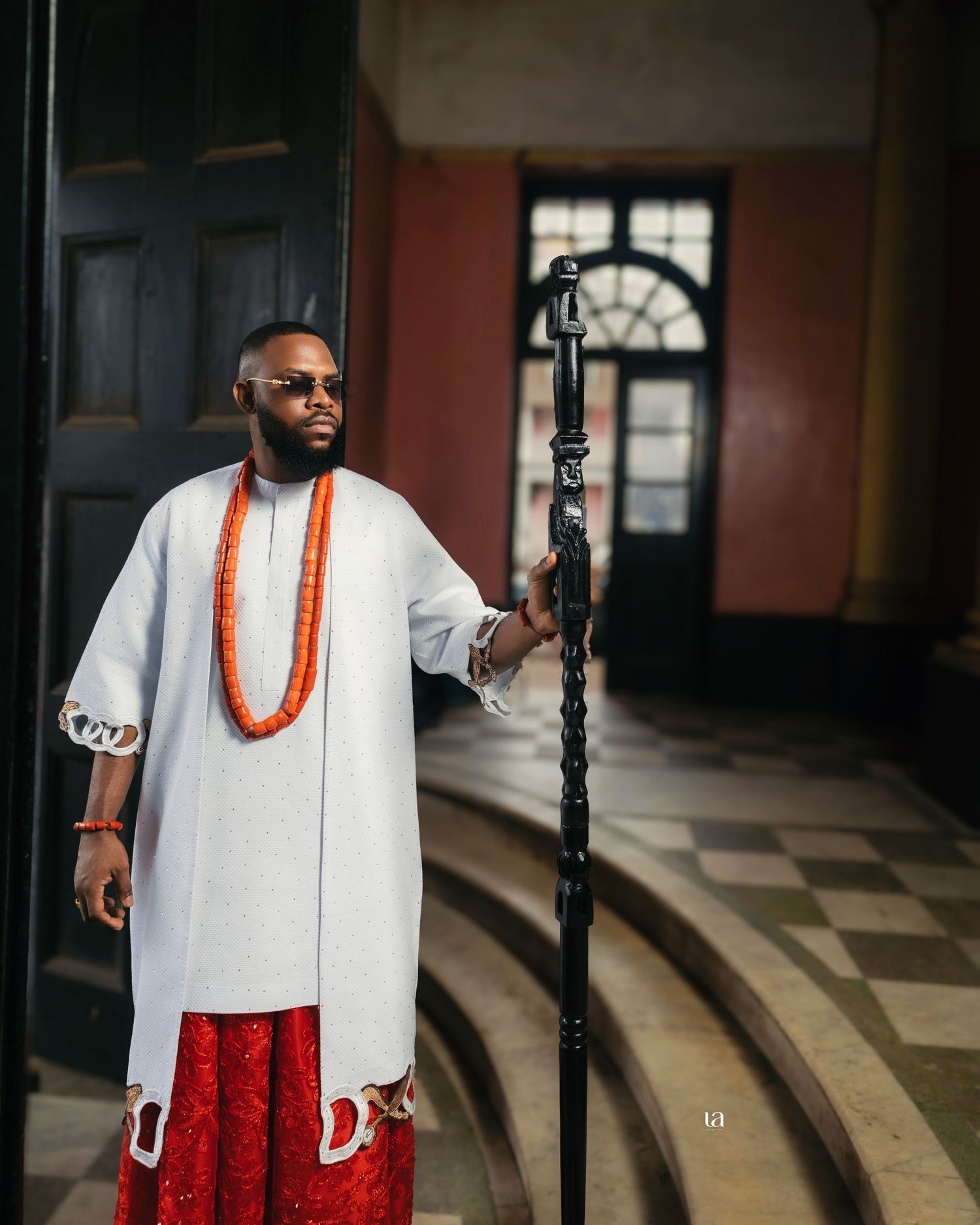 Traditional Nigerian outfit featuring white top with coral beads and red bottom in colonial architecture.