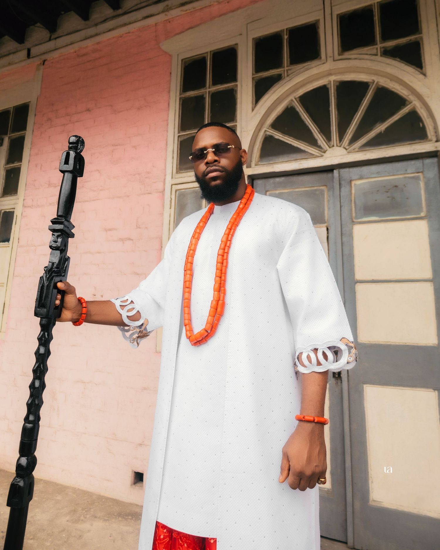 Person in white traditional attire with orange beads poses against pink wall with arched windows.