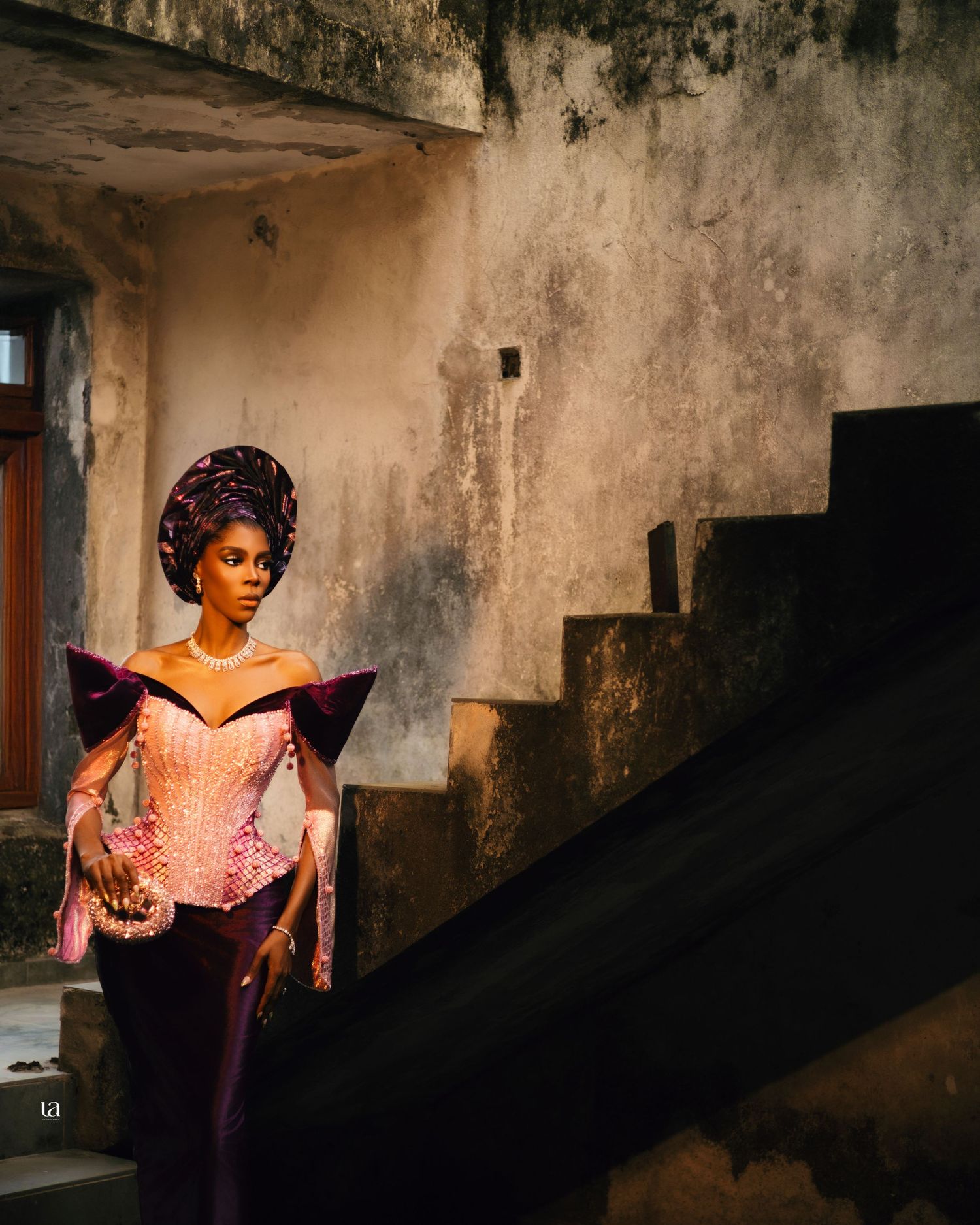 A person in a pink corset poses dramatically against a weathered wall with stone steps in the background.
