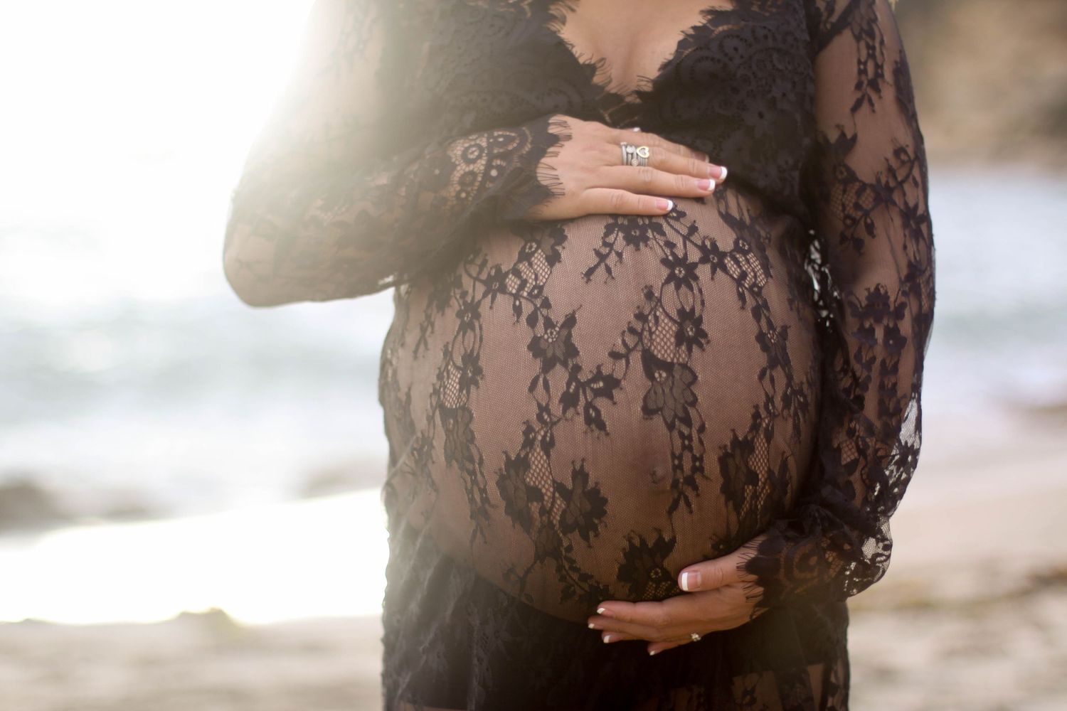 A woman in a sheer black lace dress poses elegantly against a sunlit beach background.
