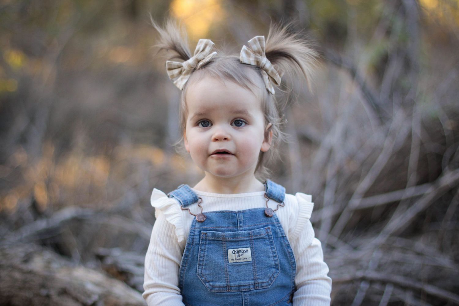 A baby in denim overalls and white shirt sits outdoors against a blurred autumn background.