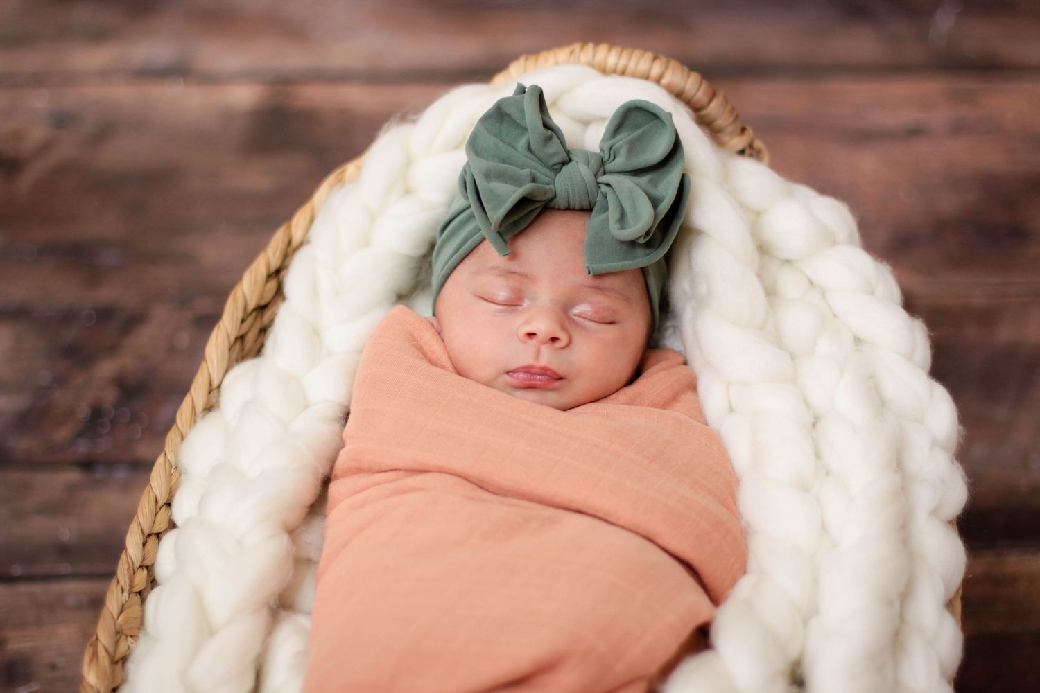 Sleeping newborn wrapped in peach swaddle with green bow in cozy basket.