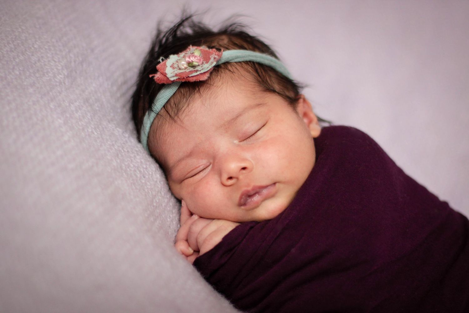 Sleeping newborn in purple wrap with floral headband against light backdrop.