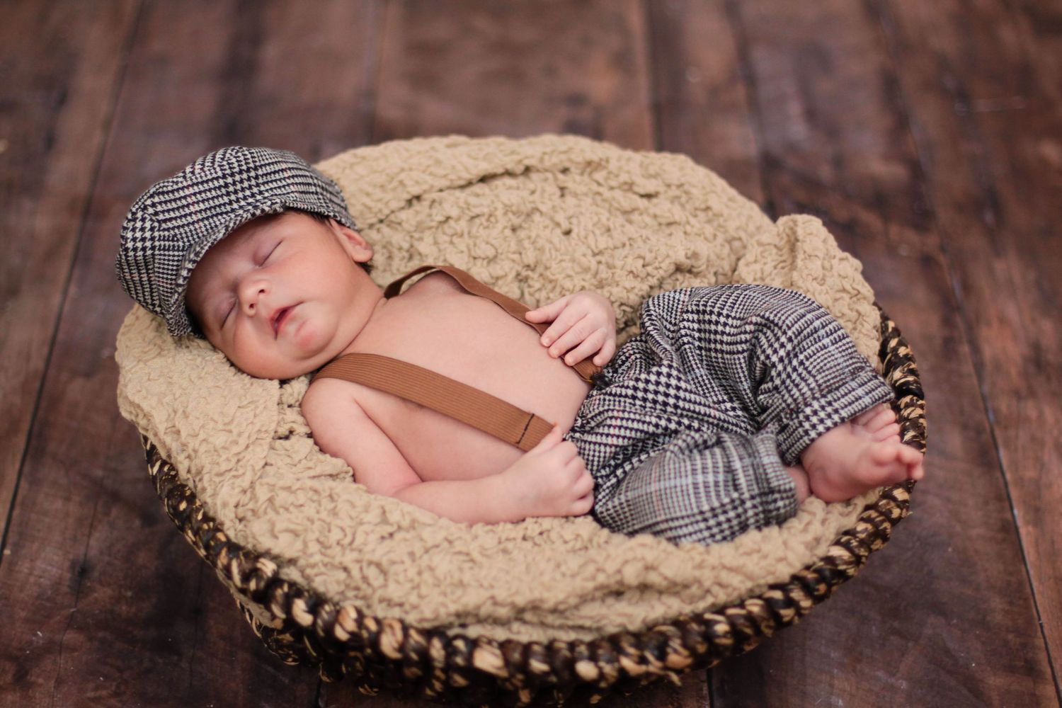 Sleeping newborn with checkered cap resting in woven basket on wooden surface.