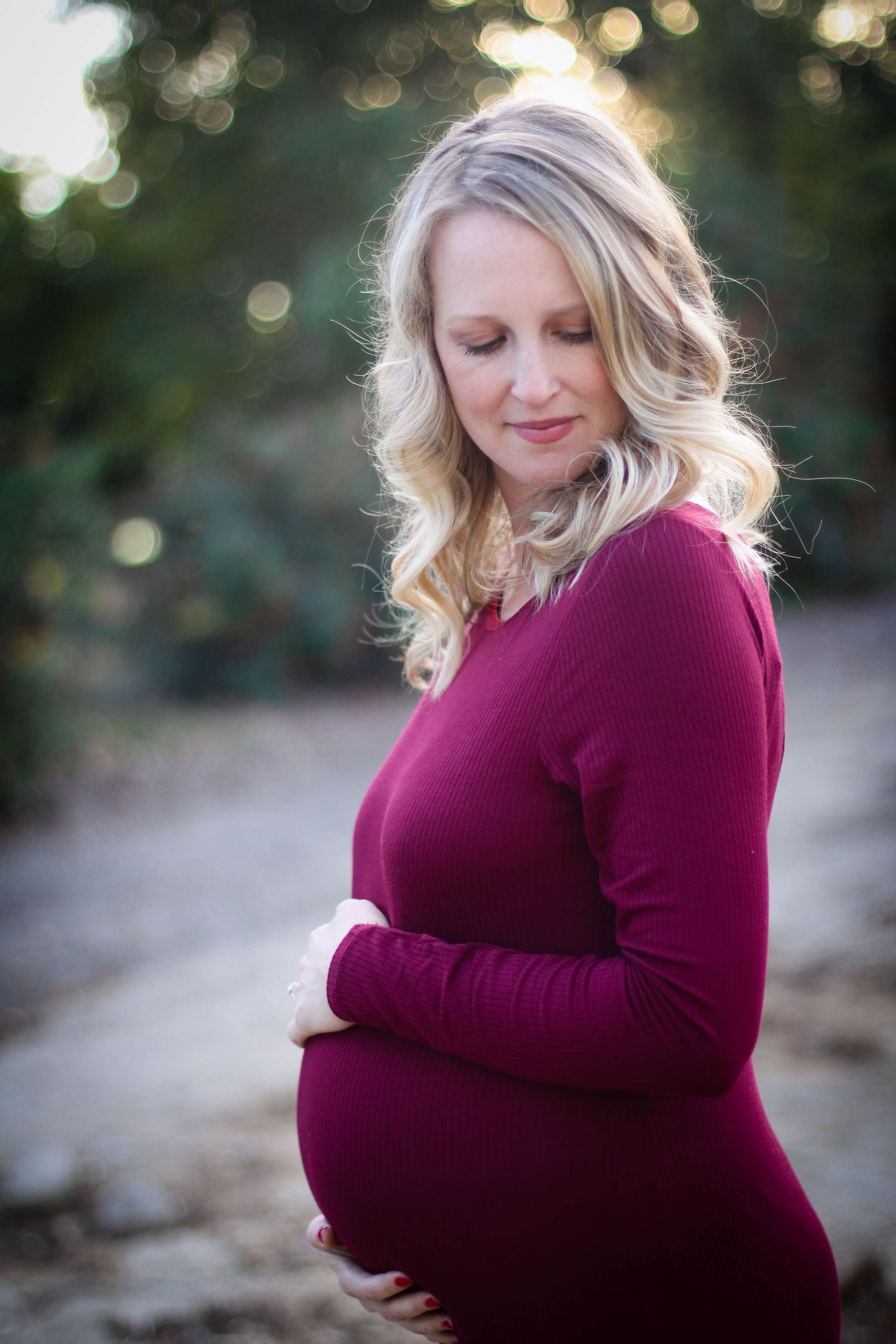 A maternity portrait outdoors shows a woman in a burgundy sweater with soft bokeh lighting in the background.