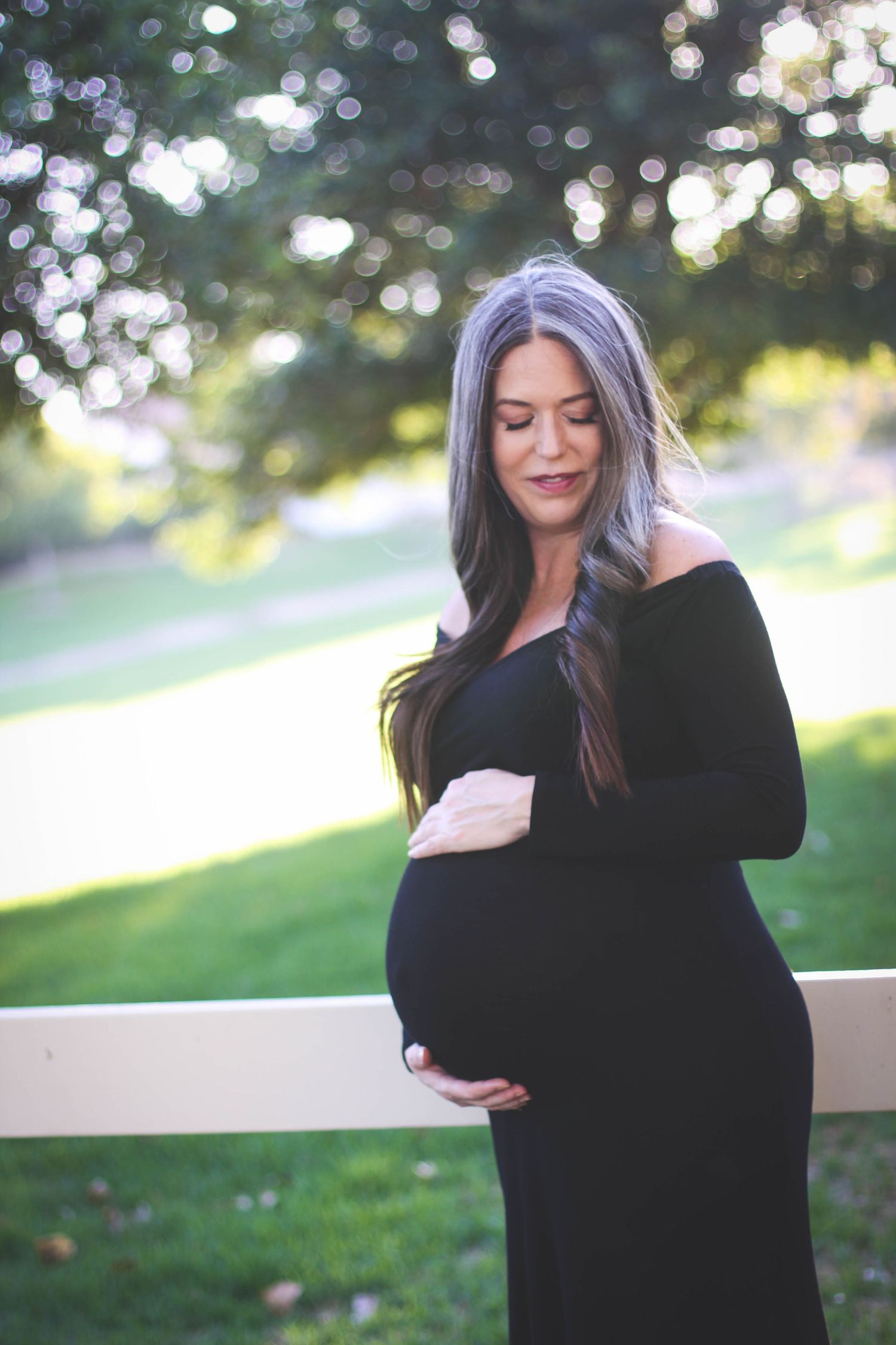 Serene maternity portrait in black off-shoulder dress against sunlit outdoor setting.