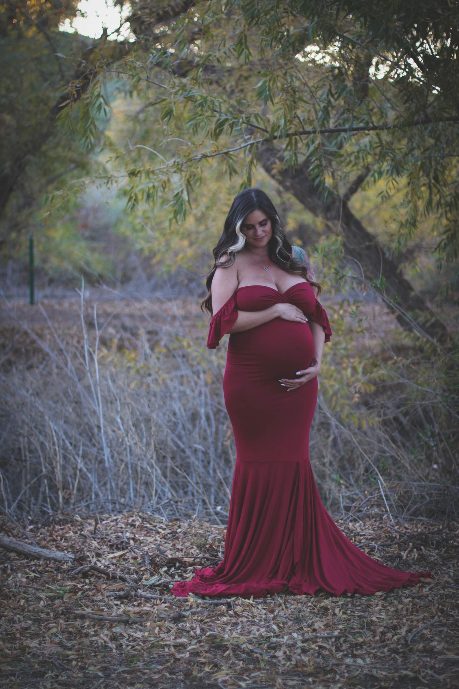 An expectant woman in a flowing burgundy gown poses gracefully in a dreamy outdoor forest setting at dusk.