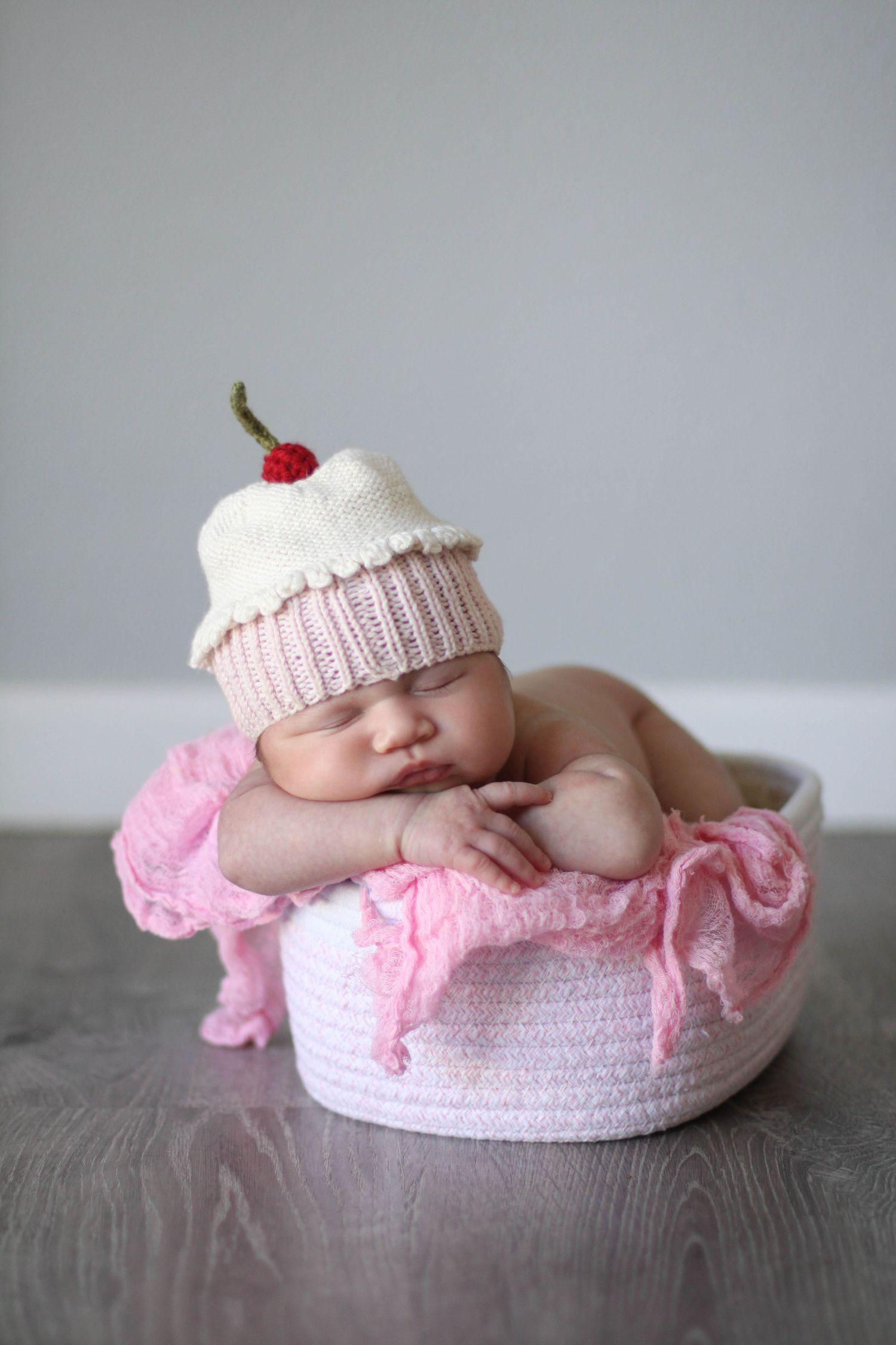 Newborn wearing white knit cupcake hat sleeps in pink cushioned basket.