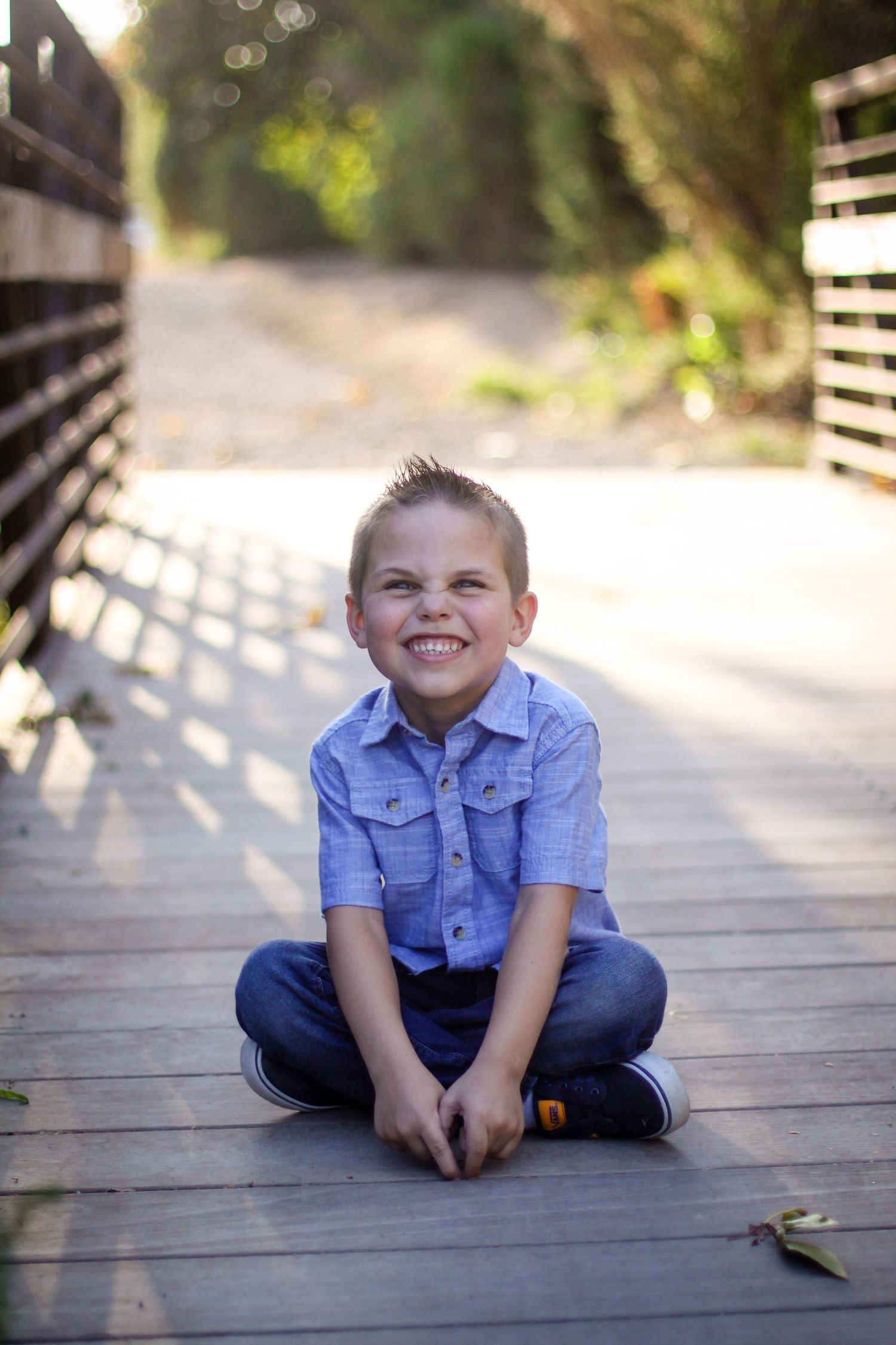 Young child in blue denim shirt sitting on wooden boardwalk for casual portrait.