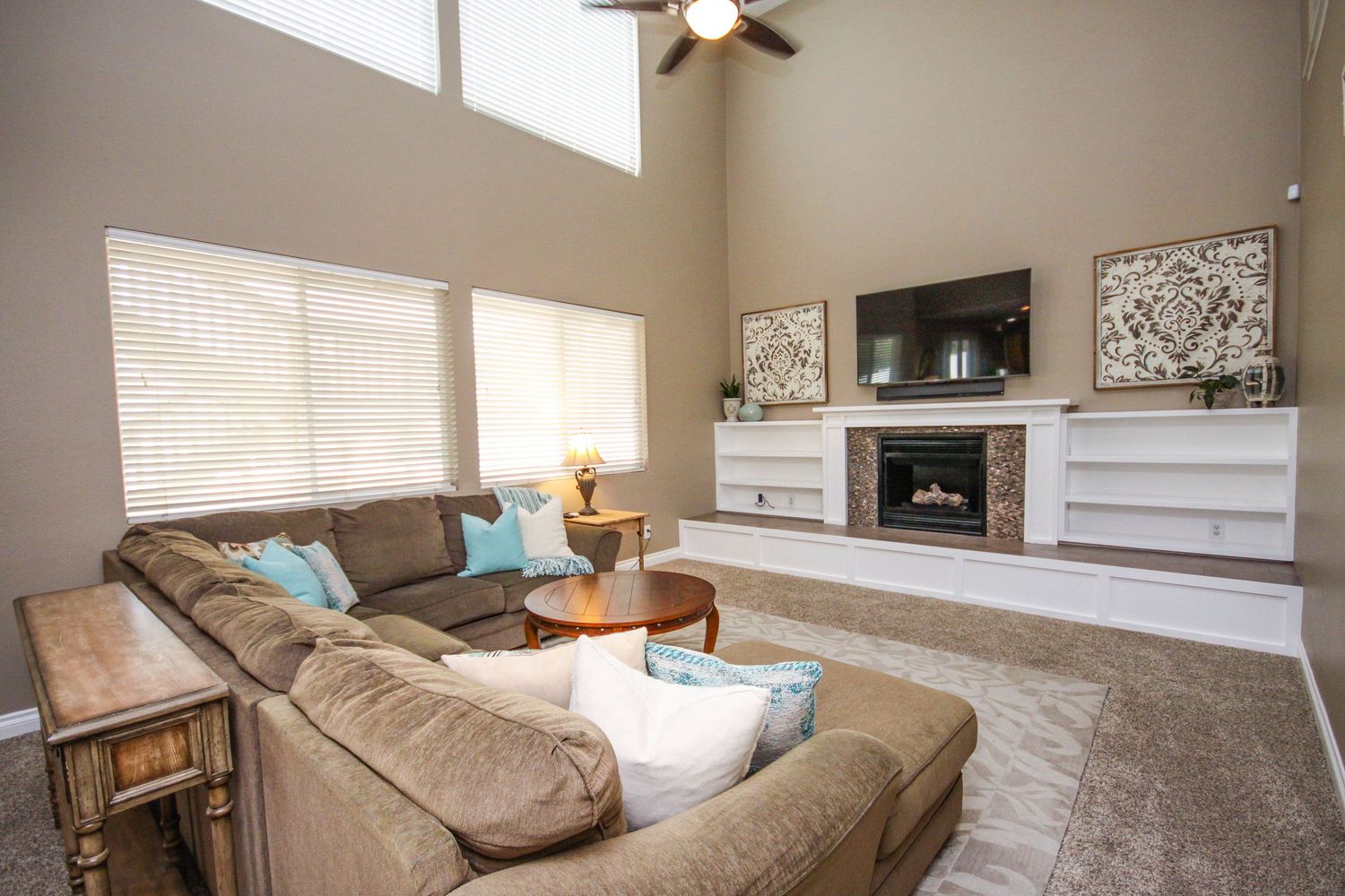 A bright and airy living room features beige carpeting, tan walls, and a ceiling fan with natural light streaming through windows.