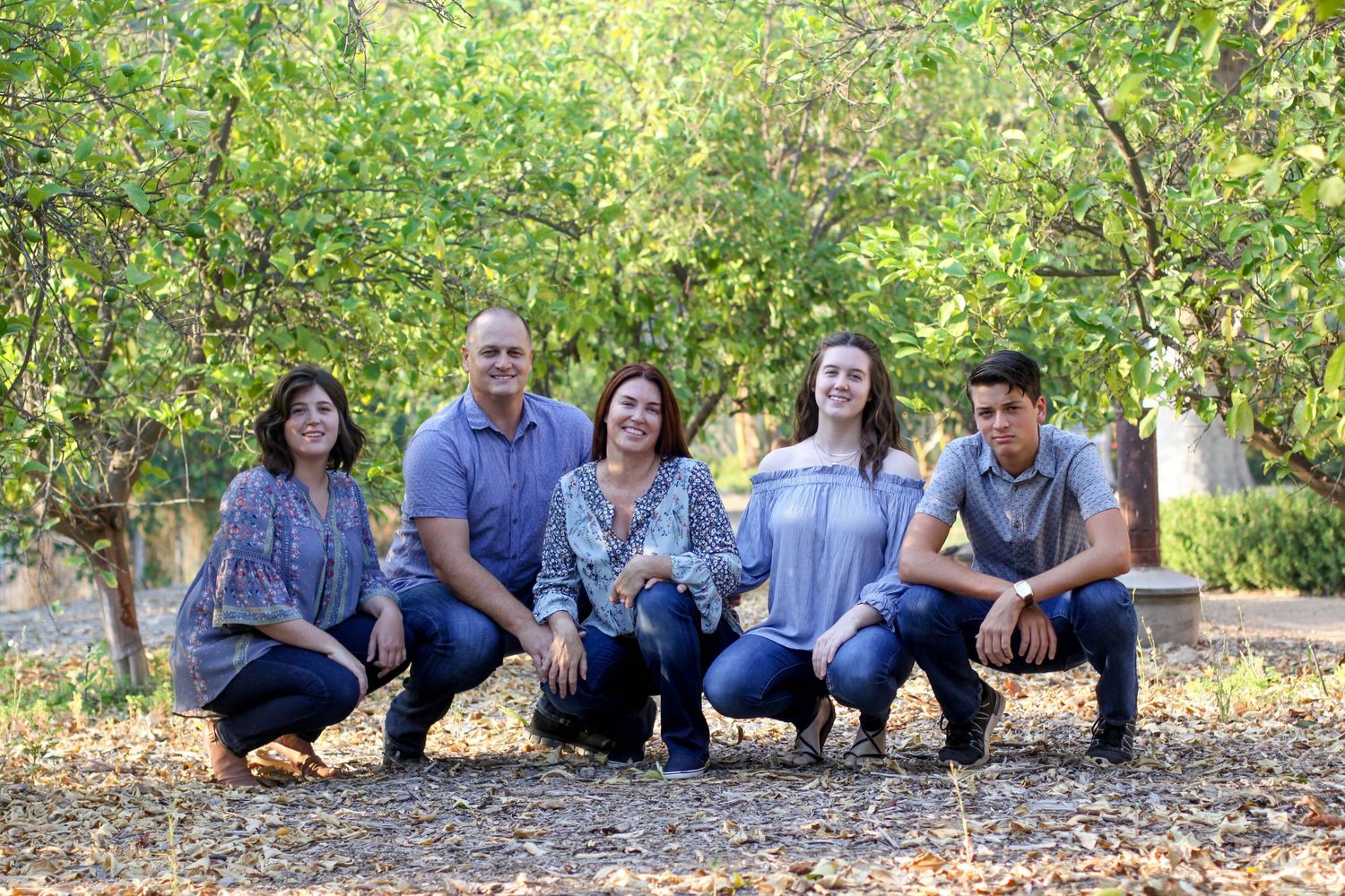 Large family group seated together on ground for informal portrait in leafy outdoor setting.