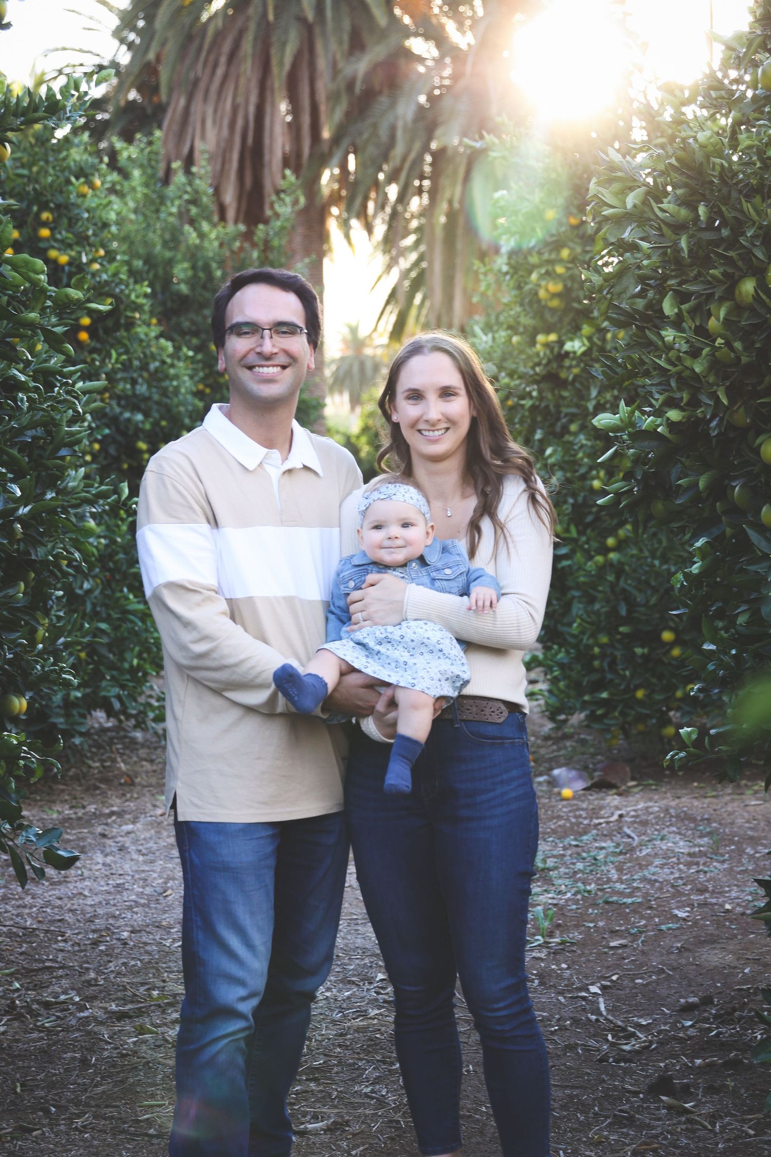 A family poses together outdoors in coordinated casual outfits with sunlight filtering through trees in the background.