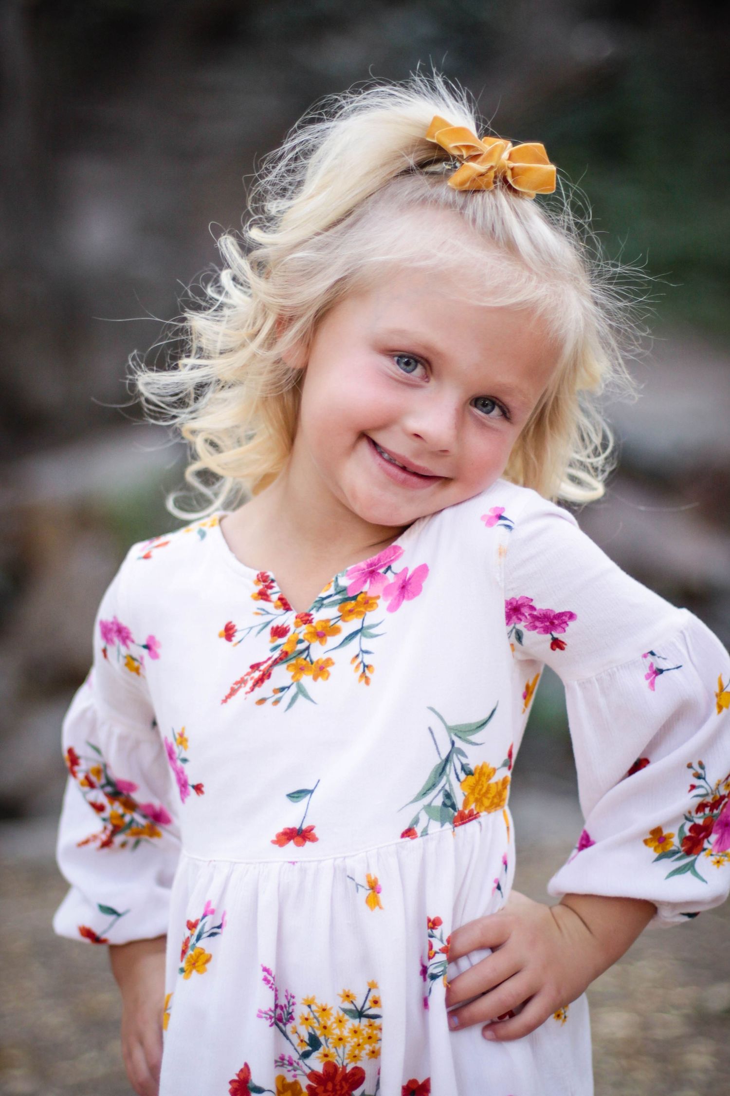 Young child in white floral dress with yellow hair bow poses playfully in a series of candid outdoor shots.
