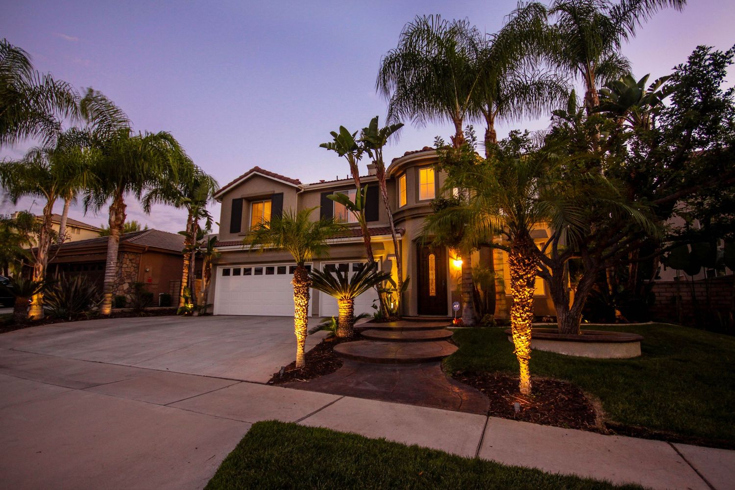 Evening view of Mediterranean home with warm landscape lighting and palm trees.