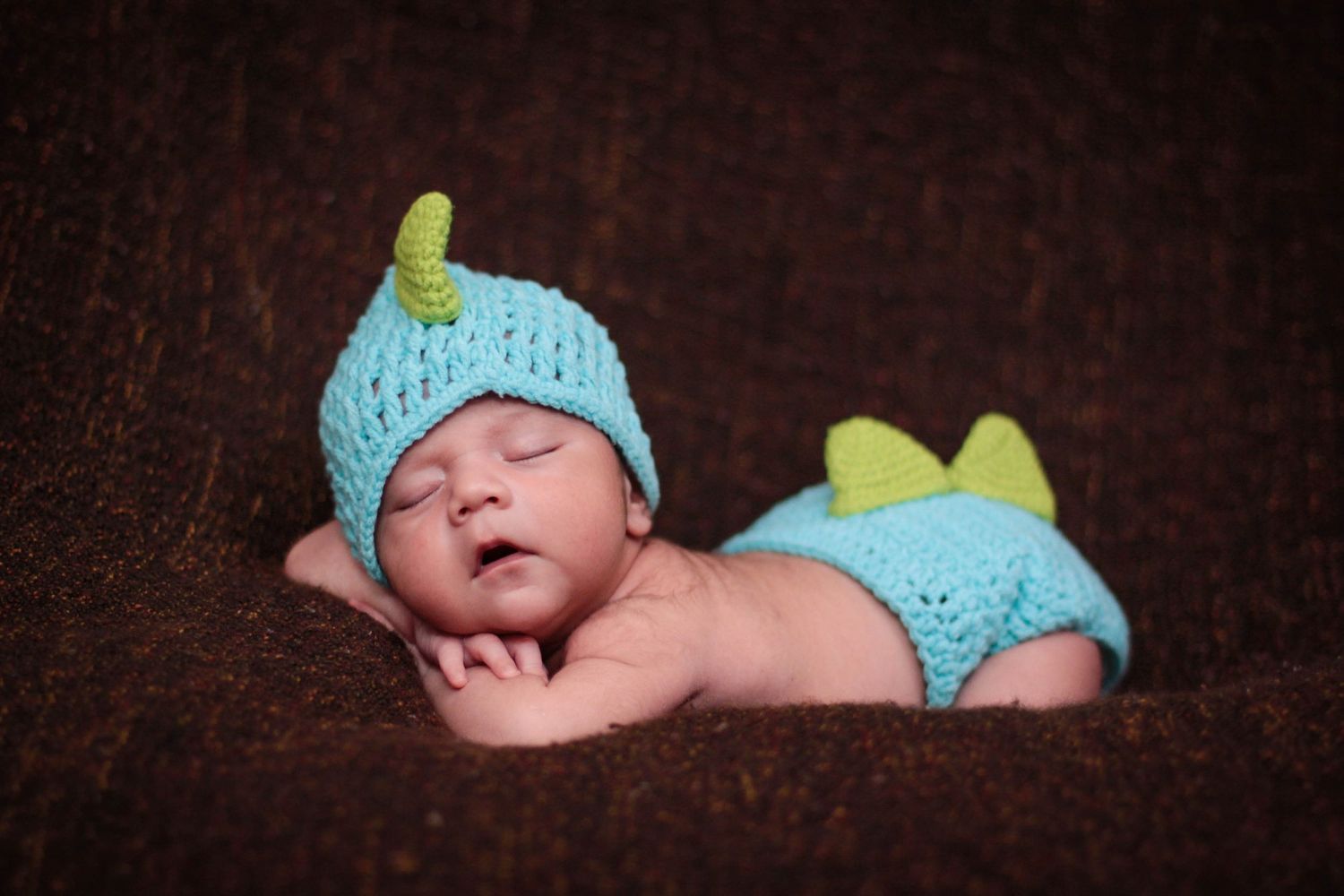 Sleeping newborn in turquoise knit outfit on dark wooden surface.