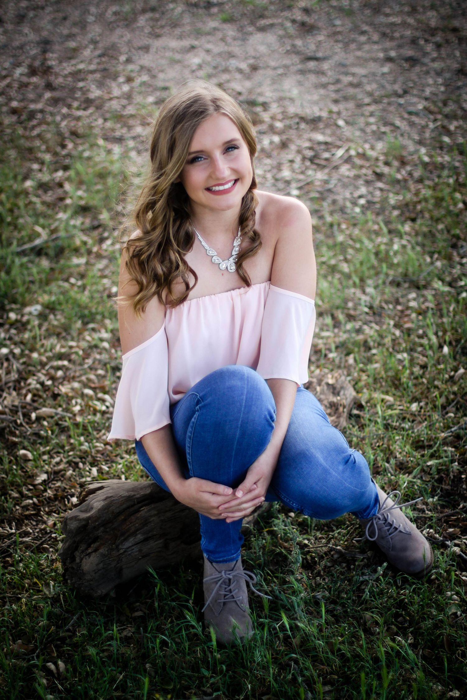 A person in an off-shoulder pink top and jeans sits on a rock in a grassy outdoor setting.